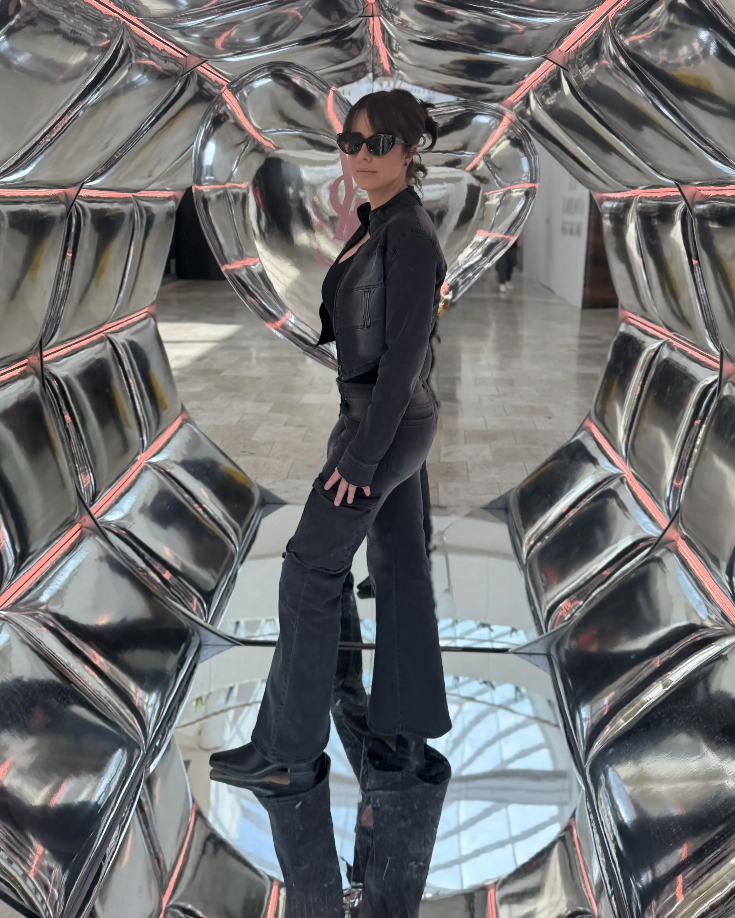 A woman wearing sunglasses, a black shirt, black pants, and a black jacket stands inside a shiny metallic balloon sculpture shaped like a heart.