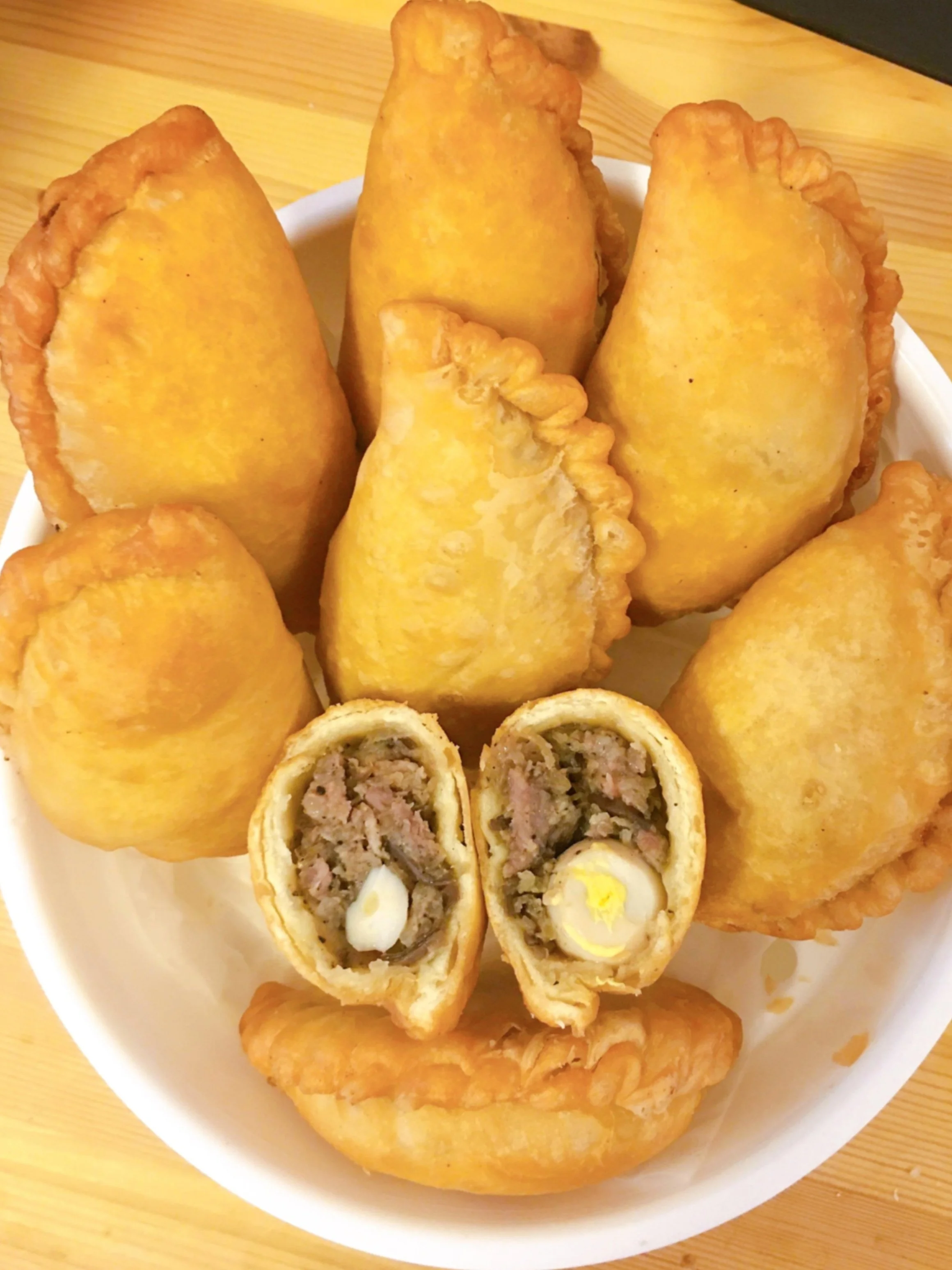 A white bowl filled with fried empanadas, one cut in half showing a meat and egg filling, placed on a wooden surface.