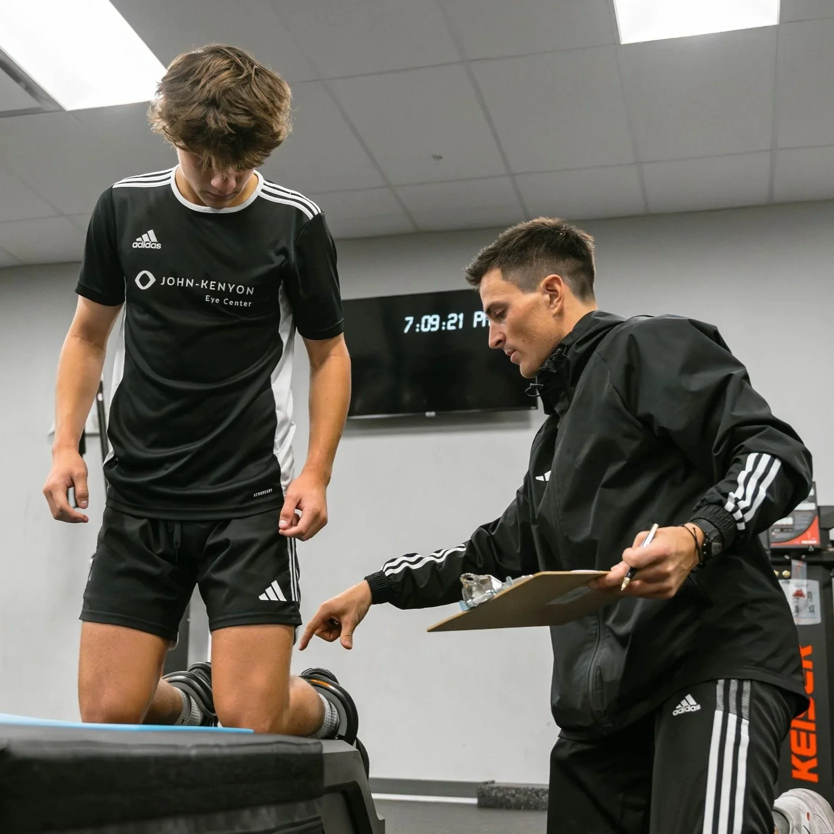 A young athlete kneeling on a treadmill during a sports performance assessment, while a coach or trainer takes notes on a clipboard.