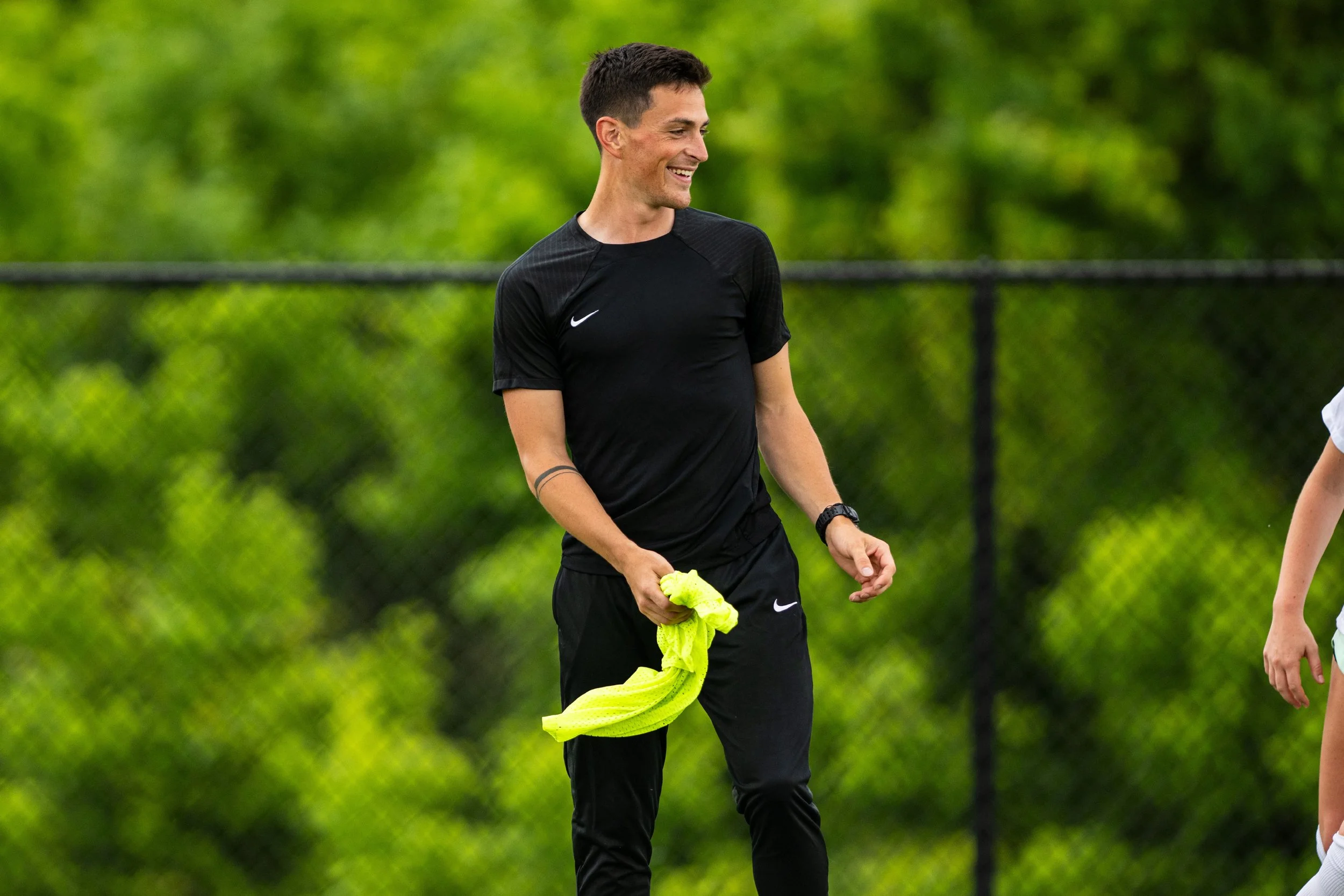 A man in black athletic wear holding a yellow shirt, smiling on a sports field with a chain-link fence and green trees in the background.