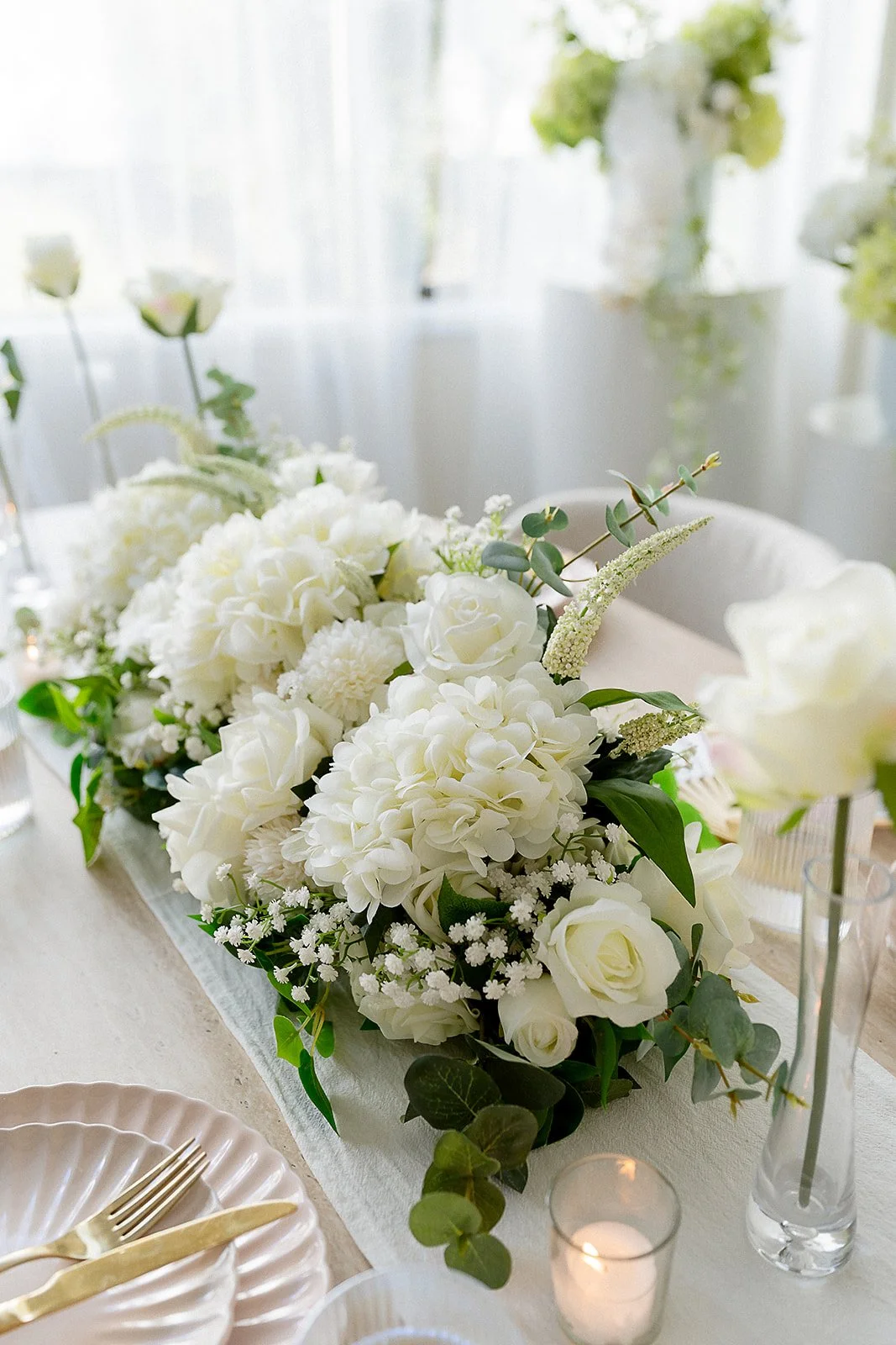 A white floral centerpiece on a dining table, featuring roses, hydrangeas, and greenery, with candles in the background and table settings including plates and cutlery.