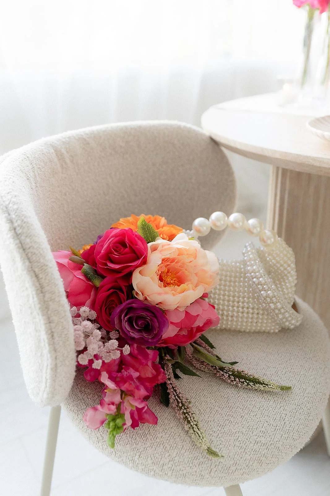 A bouquet of colorful flowers, including pink and purple roses, resting on a beige upholstered chair, with a handbag decorated with pearls nearby, in a bright room.