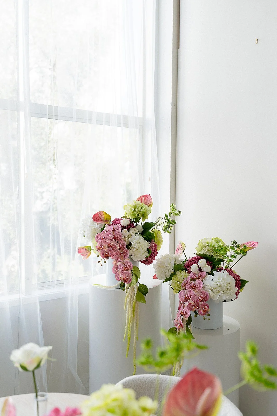Two white vases filled with pastel-colored flowers near a window with sheer white curtains in a bright room.