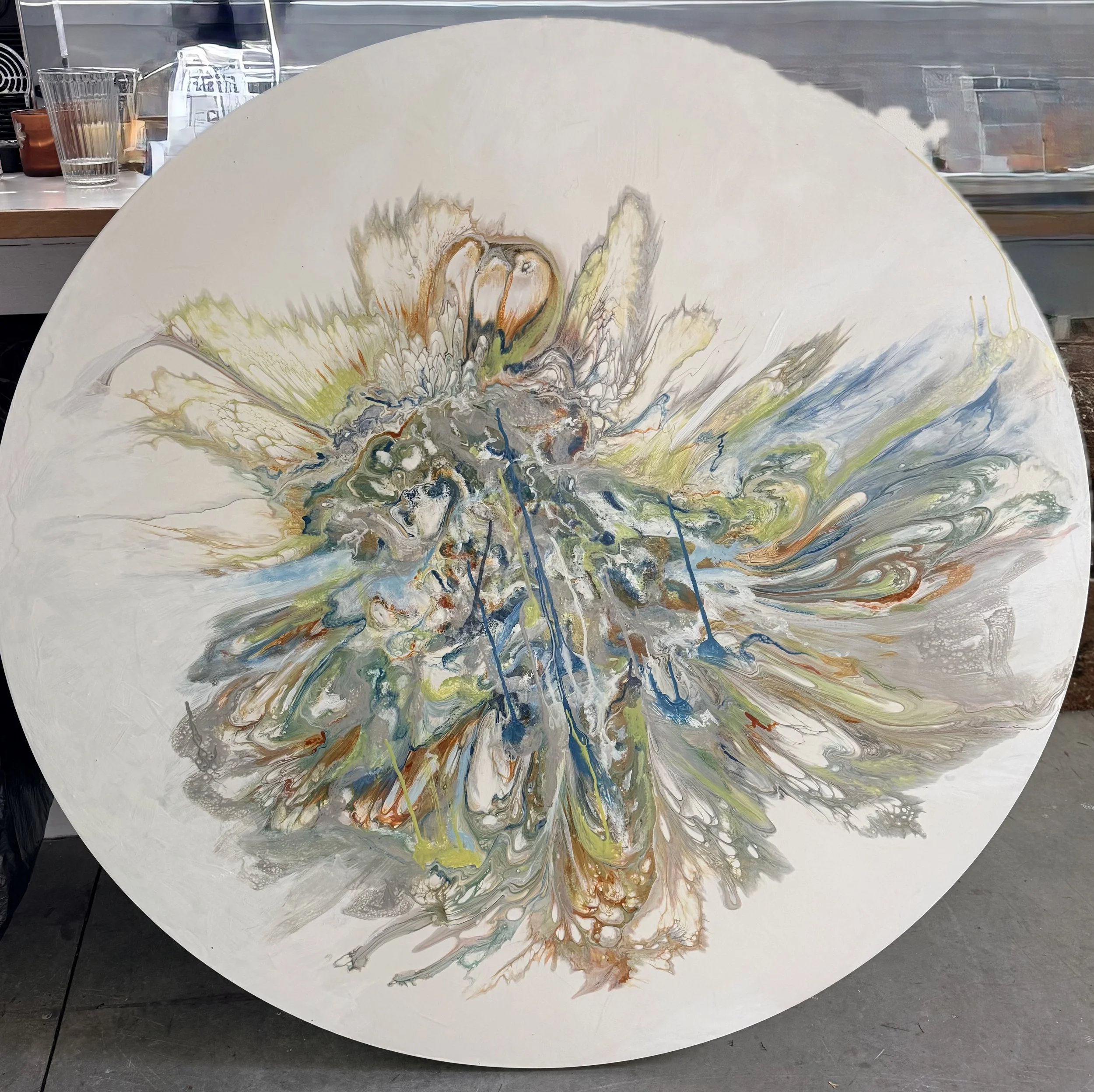 Abstract painting with swirling patterns of cream, orange, blue, green, and black paint on a large canvas. The artwork features dynamic, flowing lines radiating outward from the center.