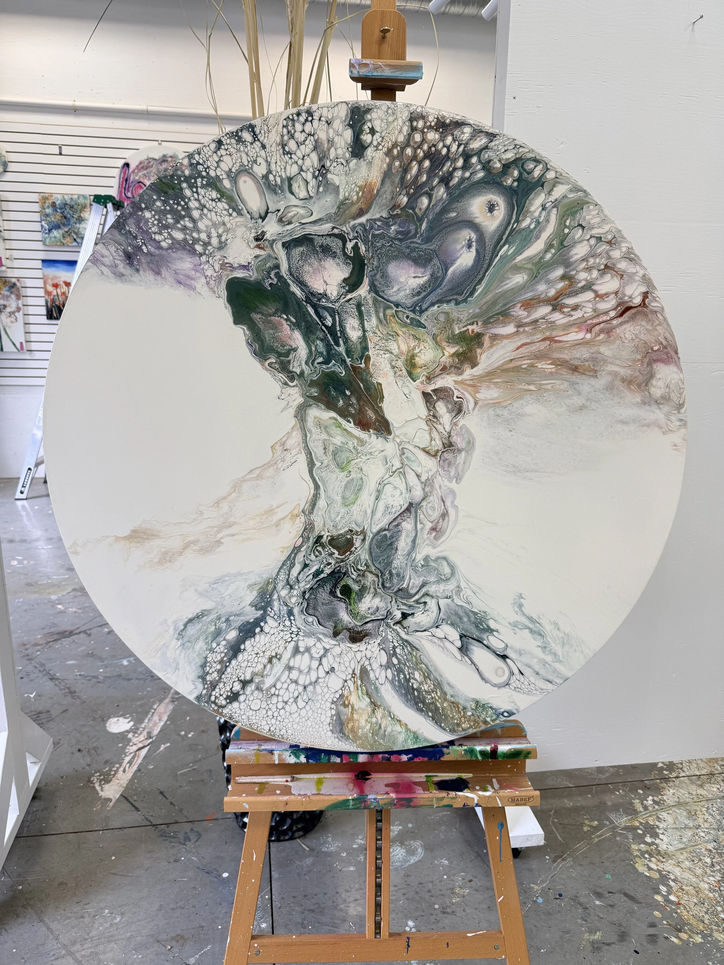 Abstract acrylic pour painting on oval canvas with swirling colors of white, black, purple, green, pink, and brown, displayed on an easel in an art studio.