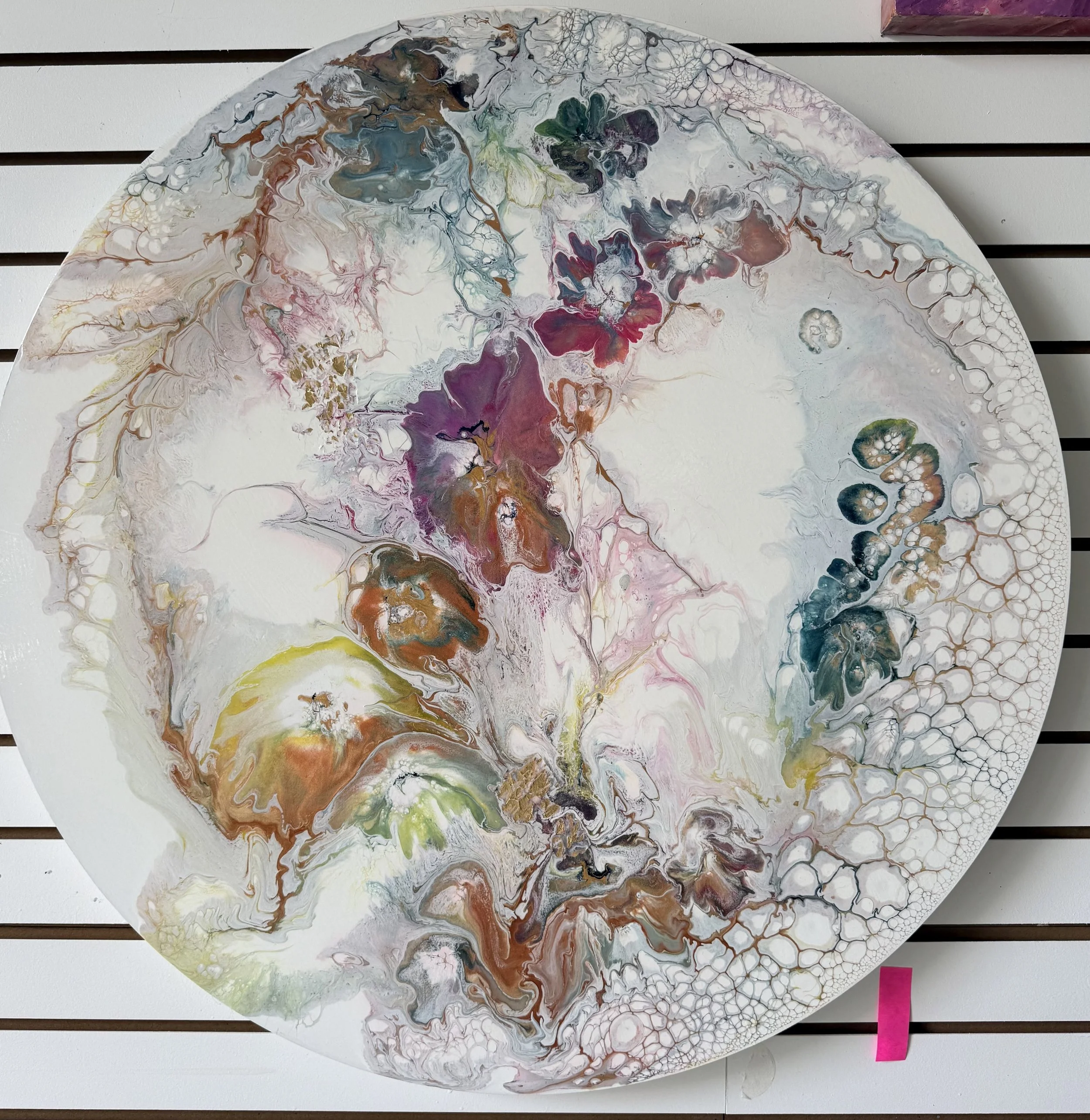 Circular abstract painting with swirling colors including white, gray, green, pink, purple, and brown, displayed against a white slatted backdrop with a pink sticky note attached.