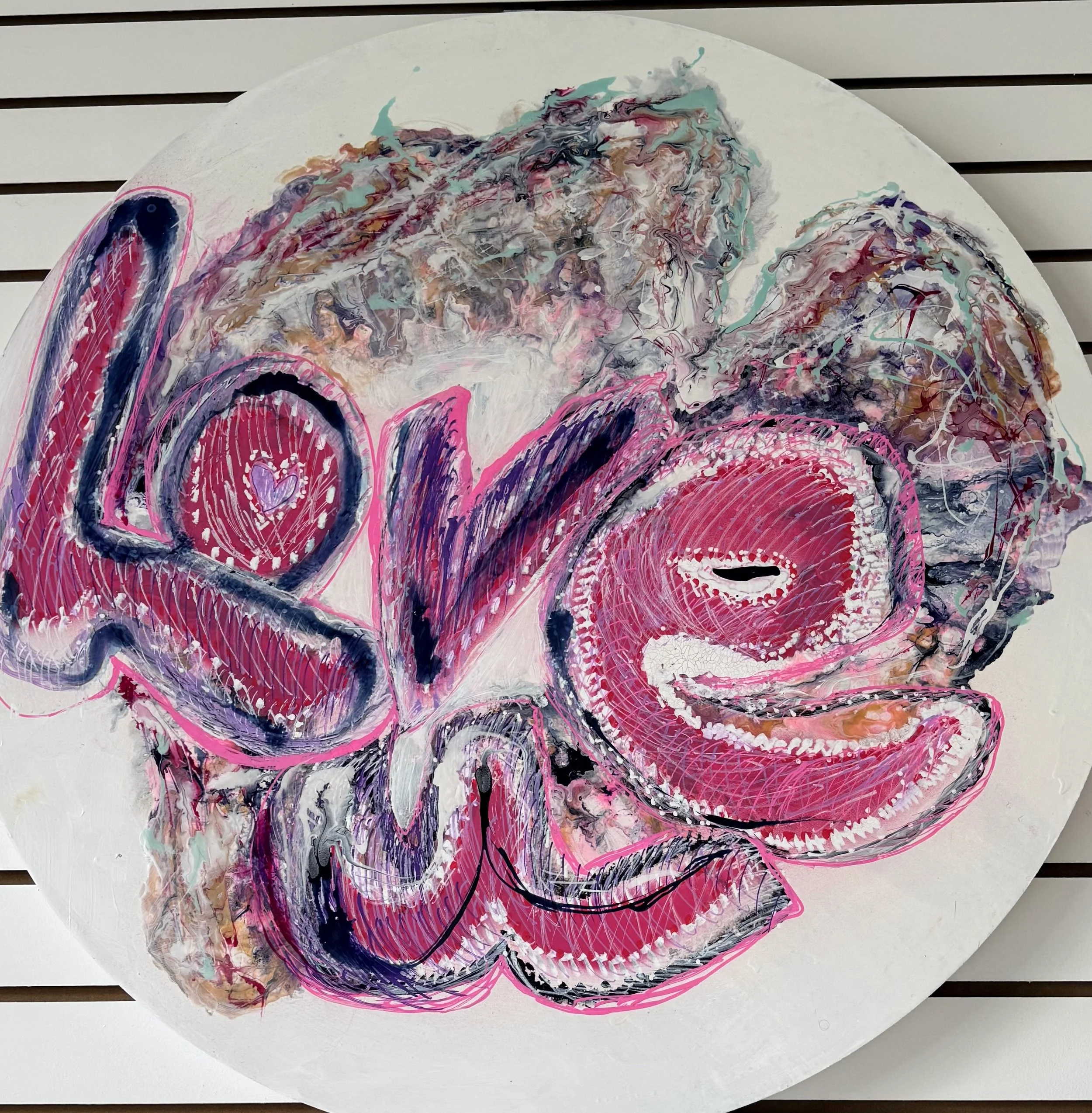 A colorful abstract painting on a circular canvas with vibrant pinks, purples, blacks, and greens, placed on a white sheet on a slatted surface.