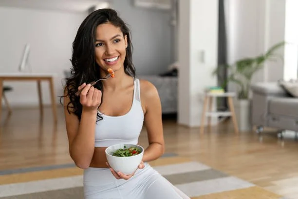 EBS: A young woman sitting on the floor in a bright living room, holding a bowl of salad and eating with a fork, smiling.