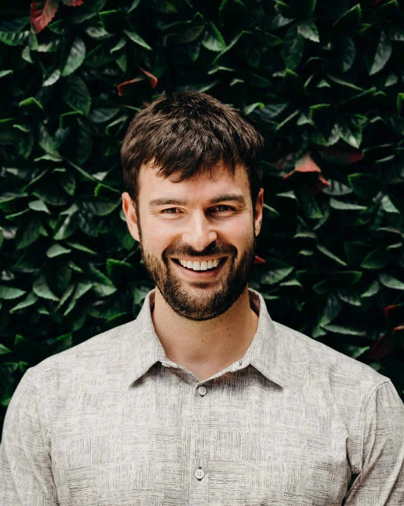 Ben - EBS Head Coach:  A man with brown hair and a beard smiling, wearing a light gray button-up shirt, standing in front of a leafy green background.