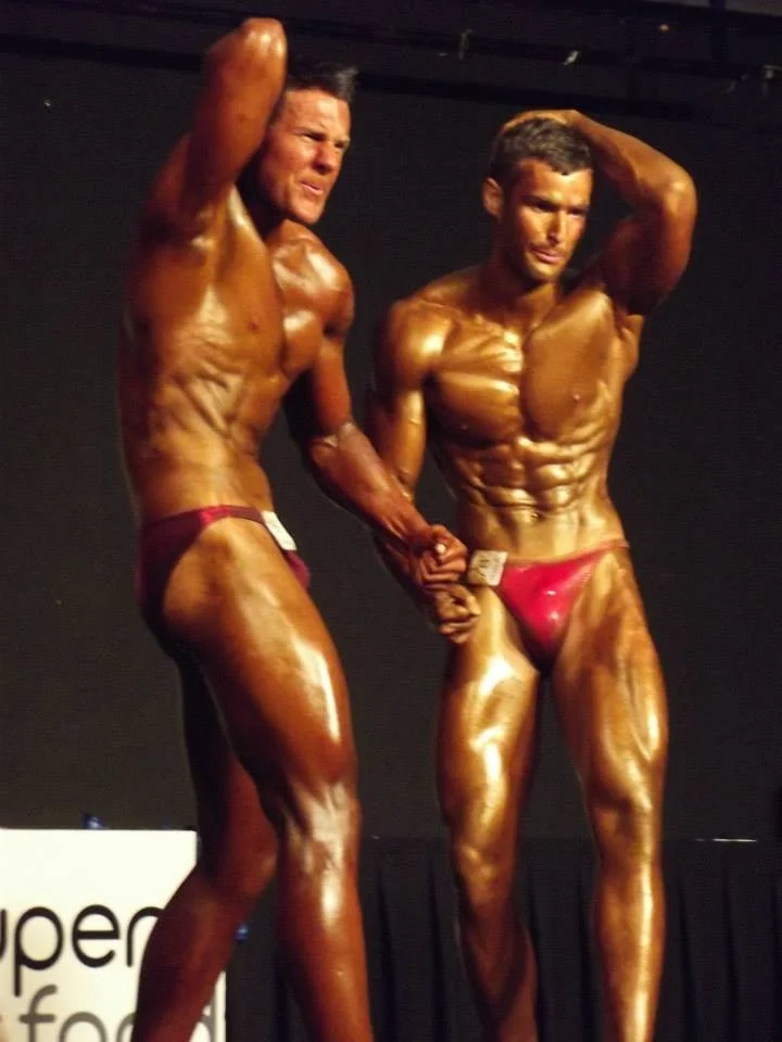 Ben - EBS Head Coach: Two muscular men in red posing trunks on stage, holding hands, during a bodybuilding competition.