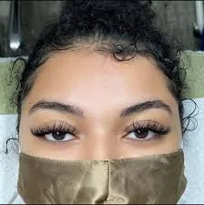 Close-up of a woman's face with curly hair and thick eyebrows, wearing a tan face mask and eyelash extensions.