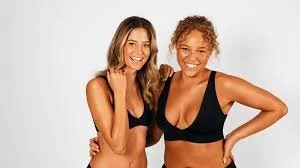 Two young women smiling, standing close together in black sports bras against a plain white background.