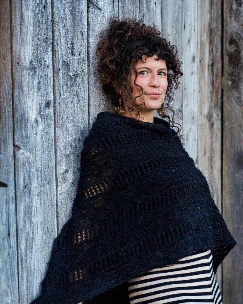 Frau mit lockigem Haar in schwarzer gestrickter Decke, die an einer Holzwand lehnt.