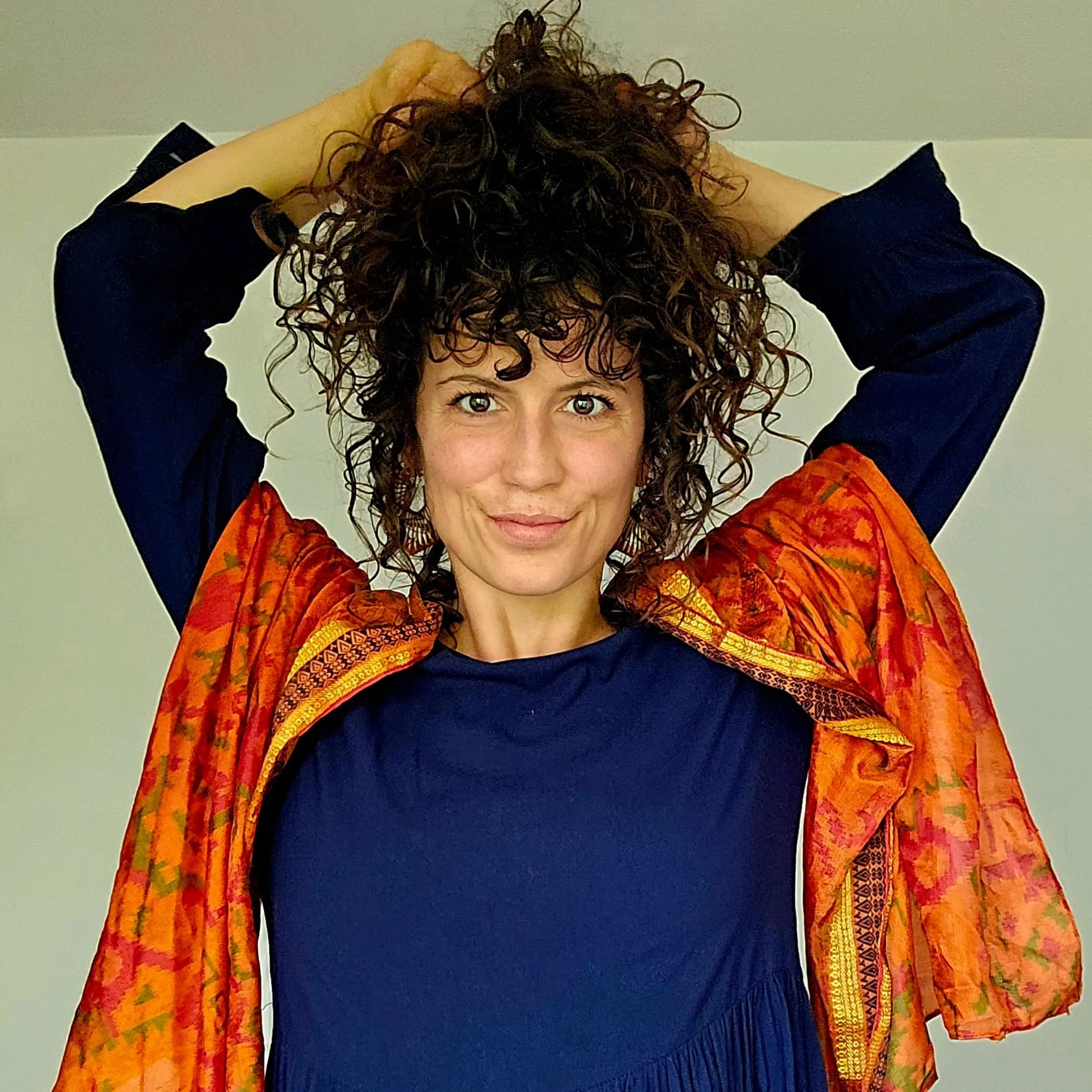 Frau mit dunkelbraunen, lockigen Haaren, die sie mit beiden Händen hält, trägt eine dunkelblaue Bluse und eine orangefarbene, bunte Jacke, steht vor hellem Hintergrund.
