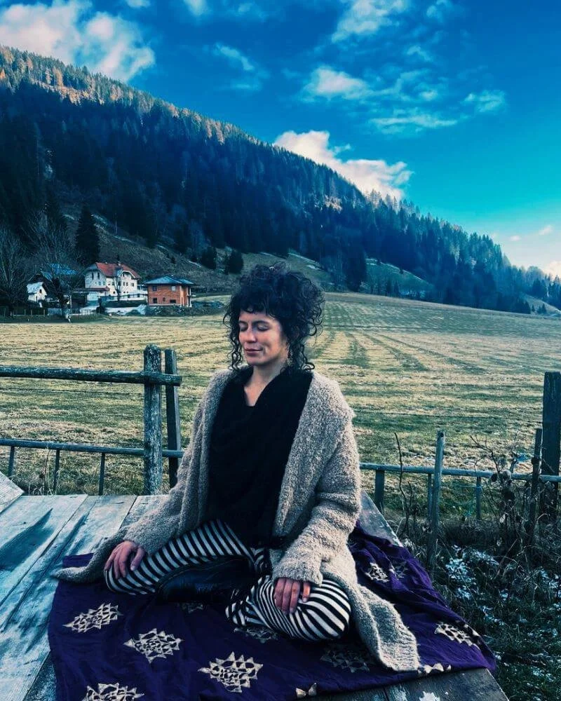 Eine Frau sitzt im Schneidersitz auf einer Decke im Freien mit einer Landschaft im Hintergrund, bestehend aus Feldern, einem Wald und Bergen unter einem blauen Himmel.