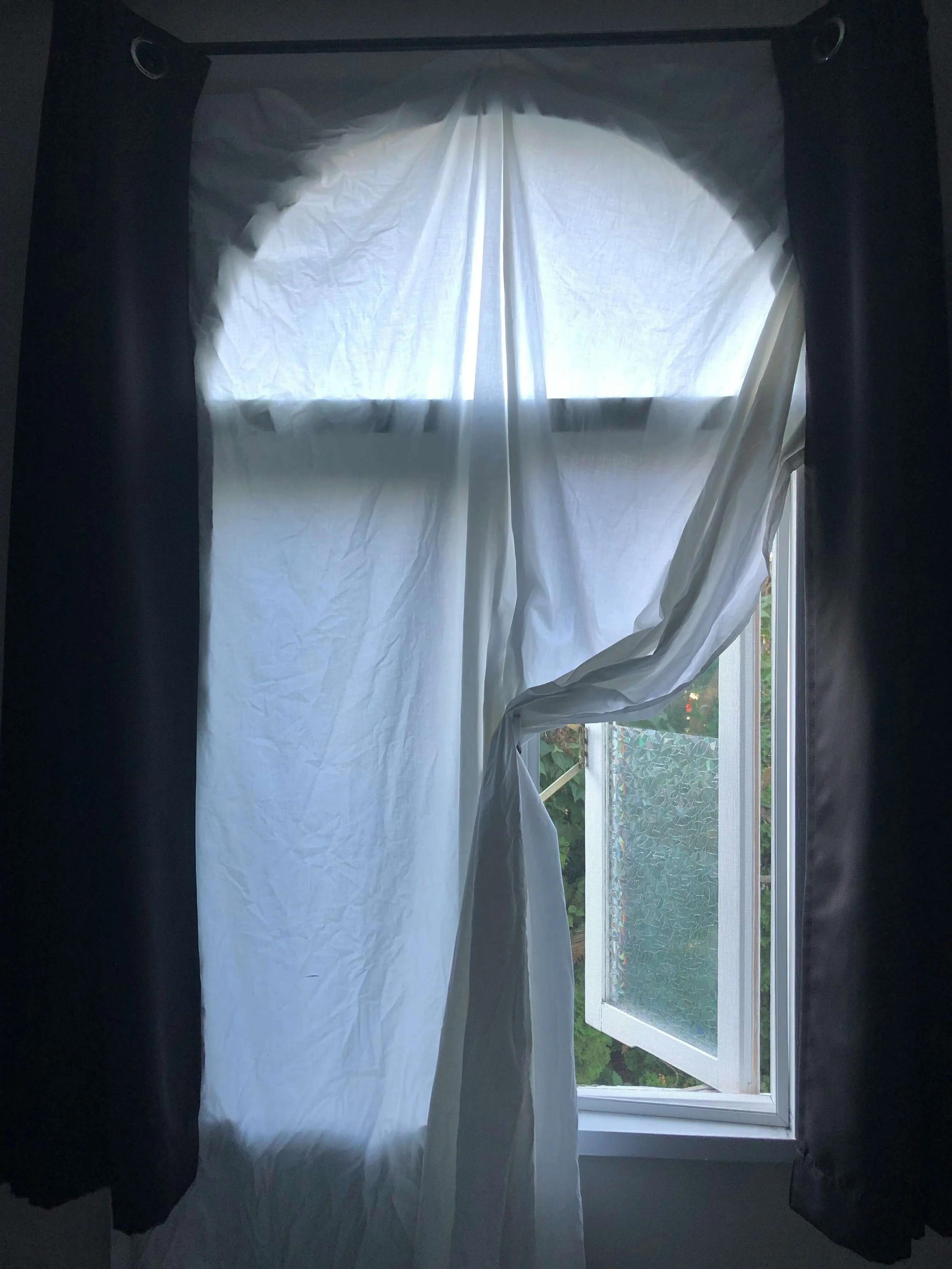 Offenes Fenster mit weißen Vorhängen, die im Wind wehen, und schwarzen Vorhängen auf beiden Seiten, Blick nach draußen auf Grünflächen.