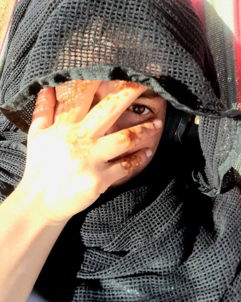 Nahaufnahme einer Person, die im Freien sitzt, mit einer schwarzen Netzjacke und einem schwarzen Kopfhörer, die die Hand vor das Gesicht hält und dabei nur ein Auge sichtbar ist.