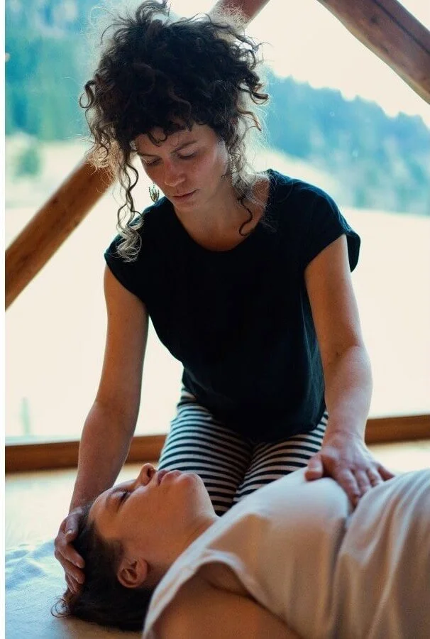 Eine Frau, die eine andere Person im Liegen behandelt, vermutlich im Rahmen einer Massage oder einer medizinischen Untersuchung. Im Hintergrund ist eine große Fensterfront mit Blick auf eine natürliche Landschaft.