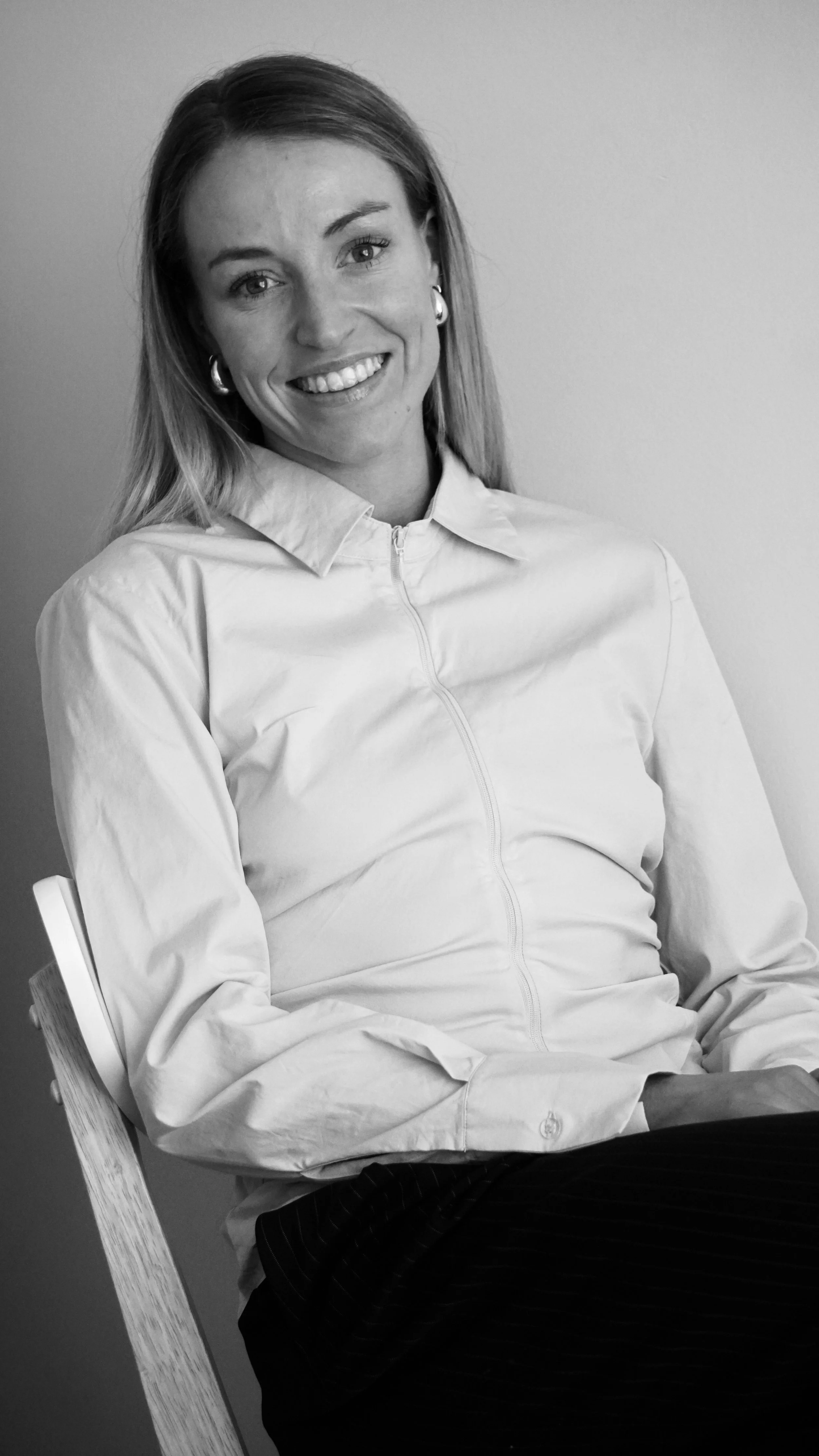 A woman smiling, sitting on a chair, wearing a zip-up collared shirt, with long hair, earrings, and a light-colored shirt in black and white.
