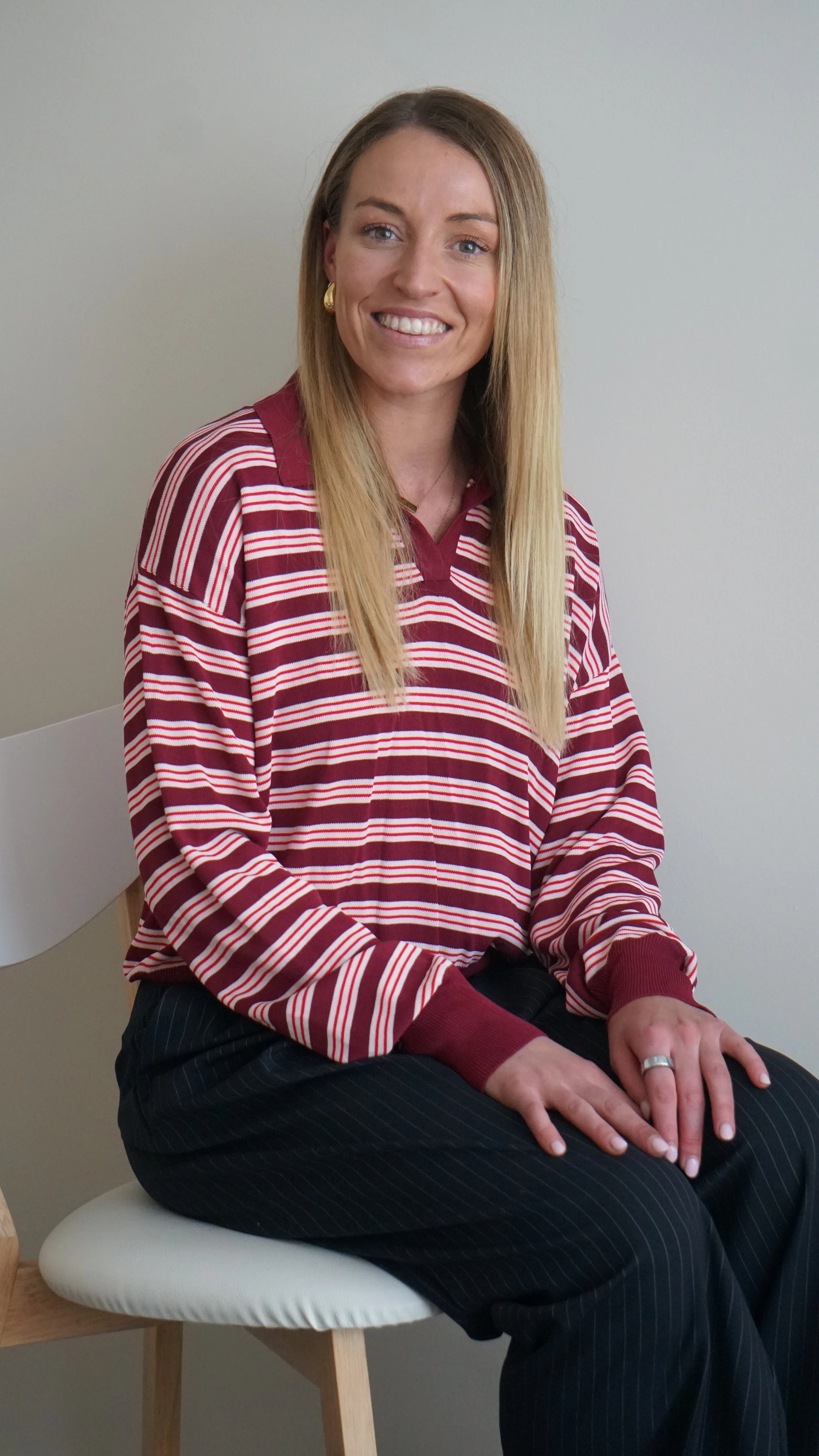 A woman with long blonde hair smiling, wearing a striped red and white Polo shirt, black pinstripe pants, and sitting on a white cushioned chair against a plain wall.