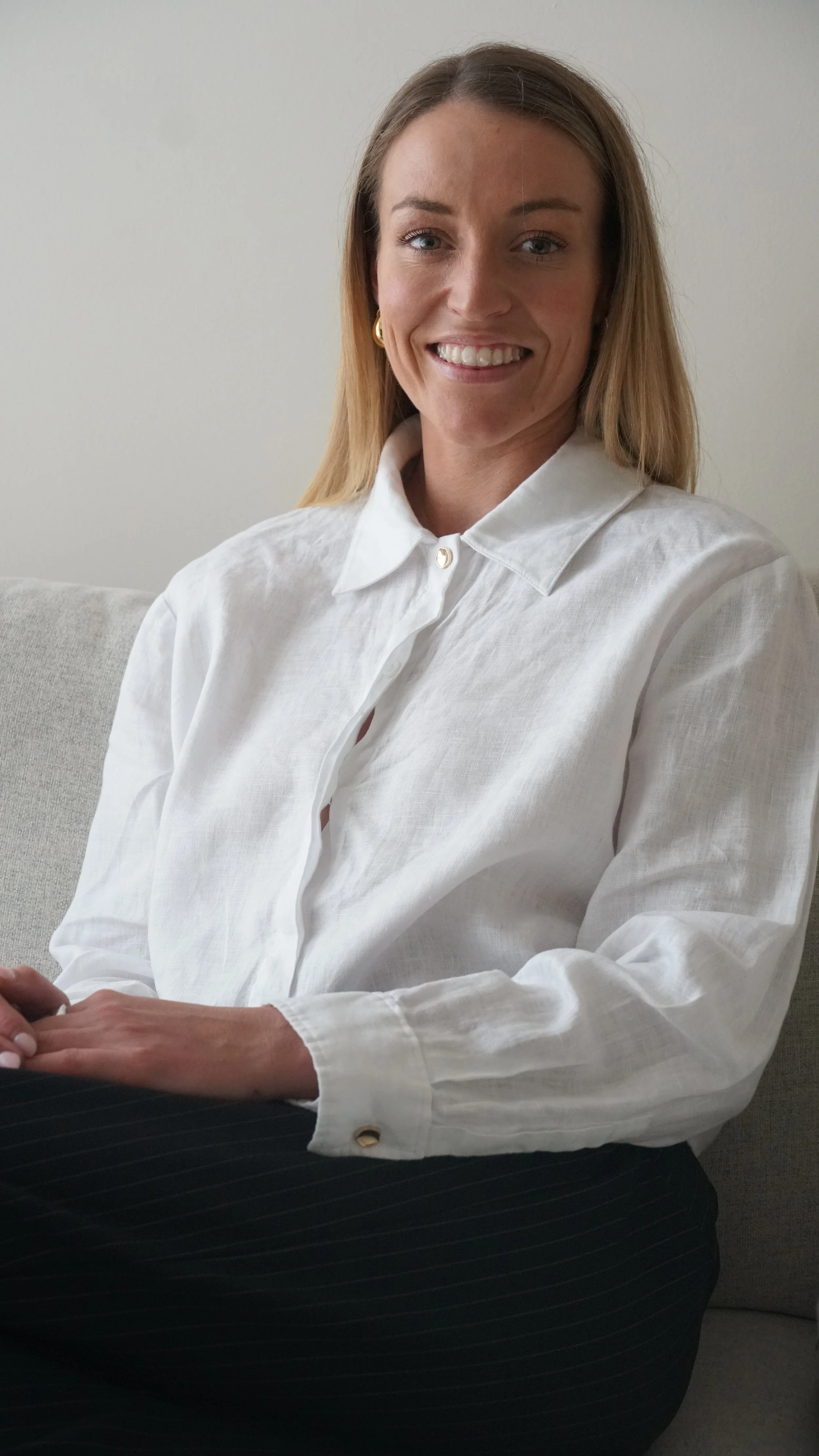 A woman sitting on a light-colored sofa, smiling at the camera, wearing a white button-up shirt and gold earrings.