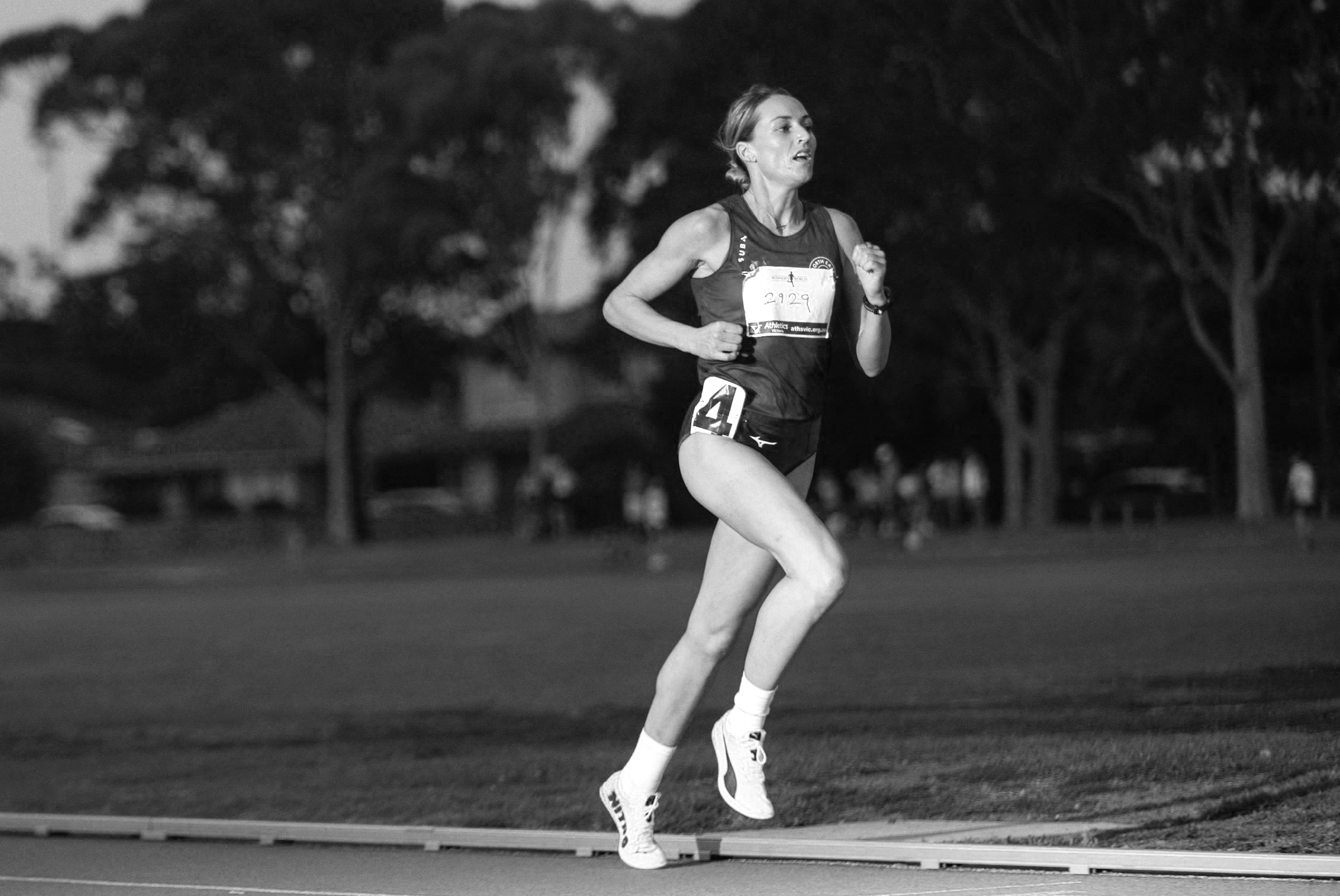 A female runner in a race, running on a track with trees and spectators in the background, in black and white.