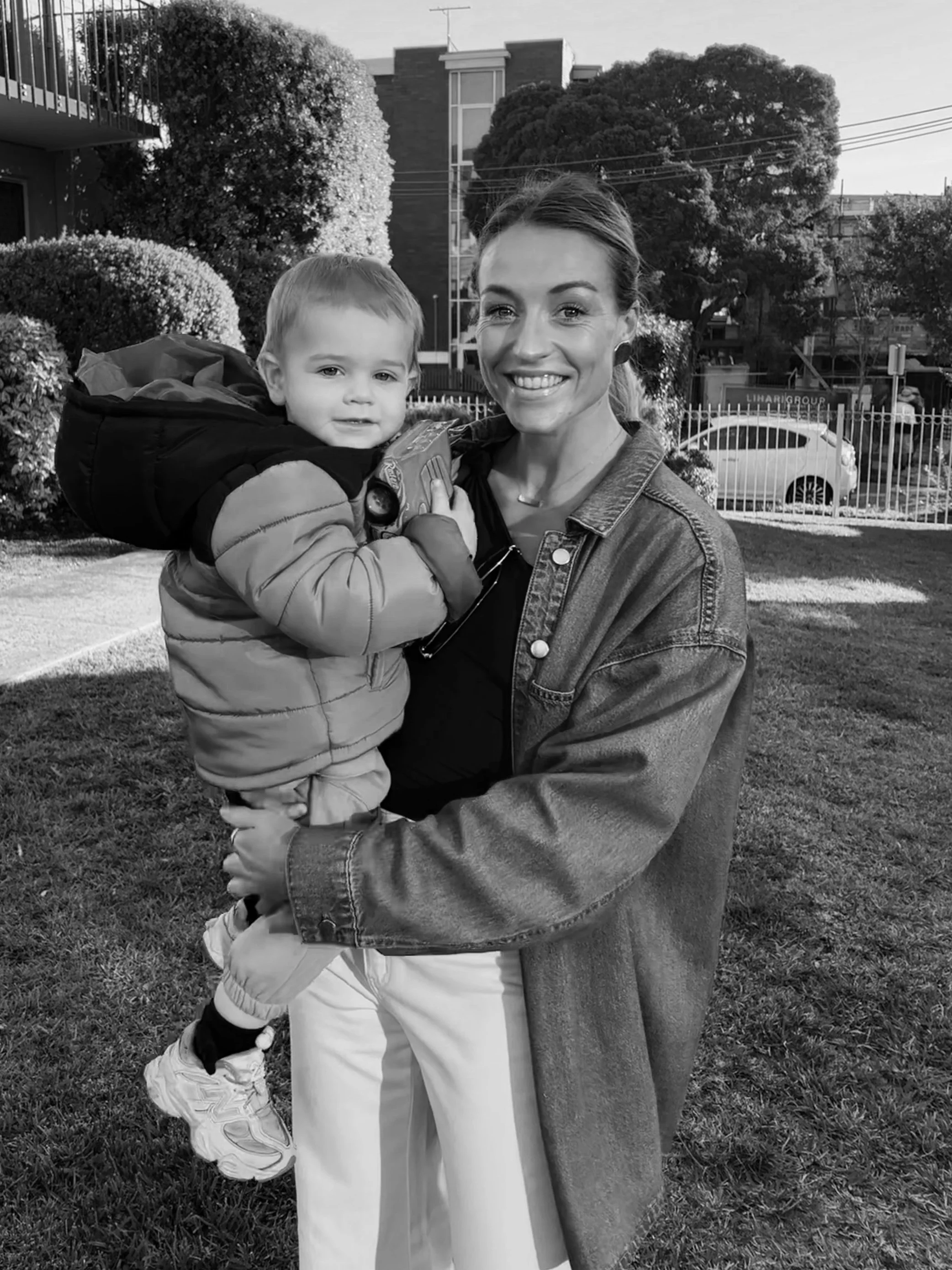 A woman holding a young boy outdoors in a park or yard, smiling at the camera with trees and buildings in the background.