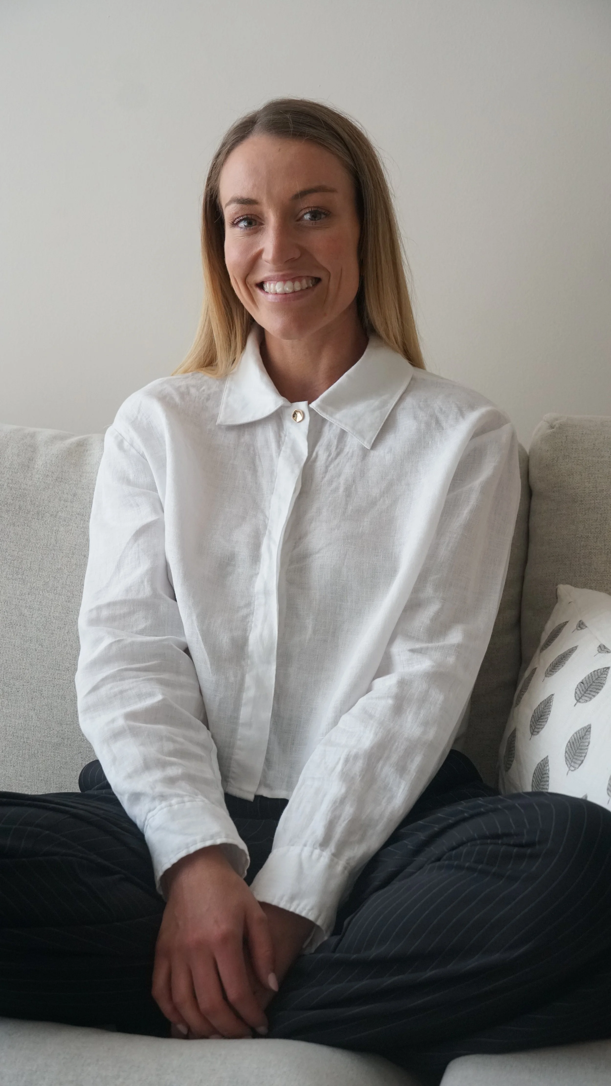 A woman with blonde hair sitting cross-legged on a light-colored sofa, smiling at the camera, wearing a white button-up shirt and dark pinstriped pants, in a room with a plain light wall and a patterned pillow.