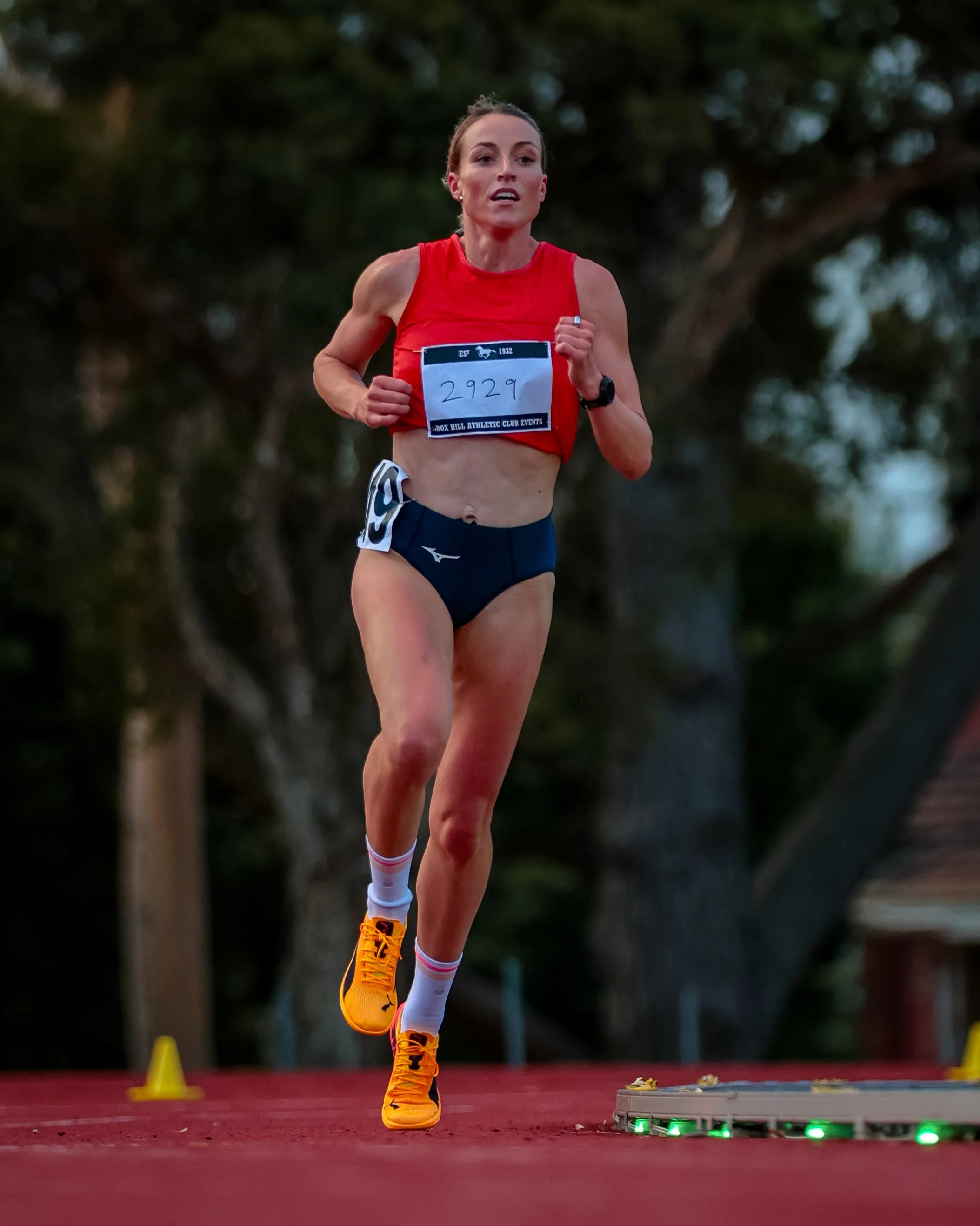 An athletic woman running on a track with a tree-lined background, wearing a red top, black shorts, orange running shoes, and a bib number 2929.