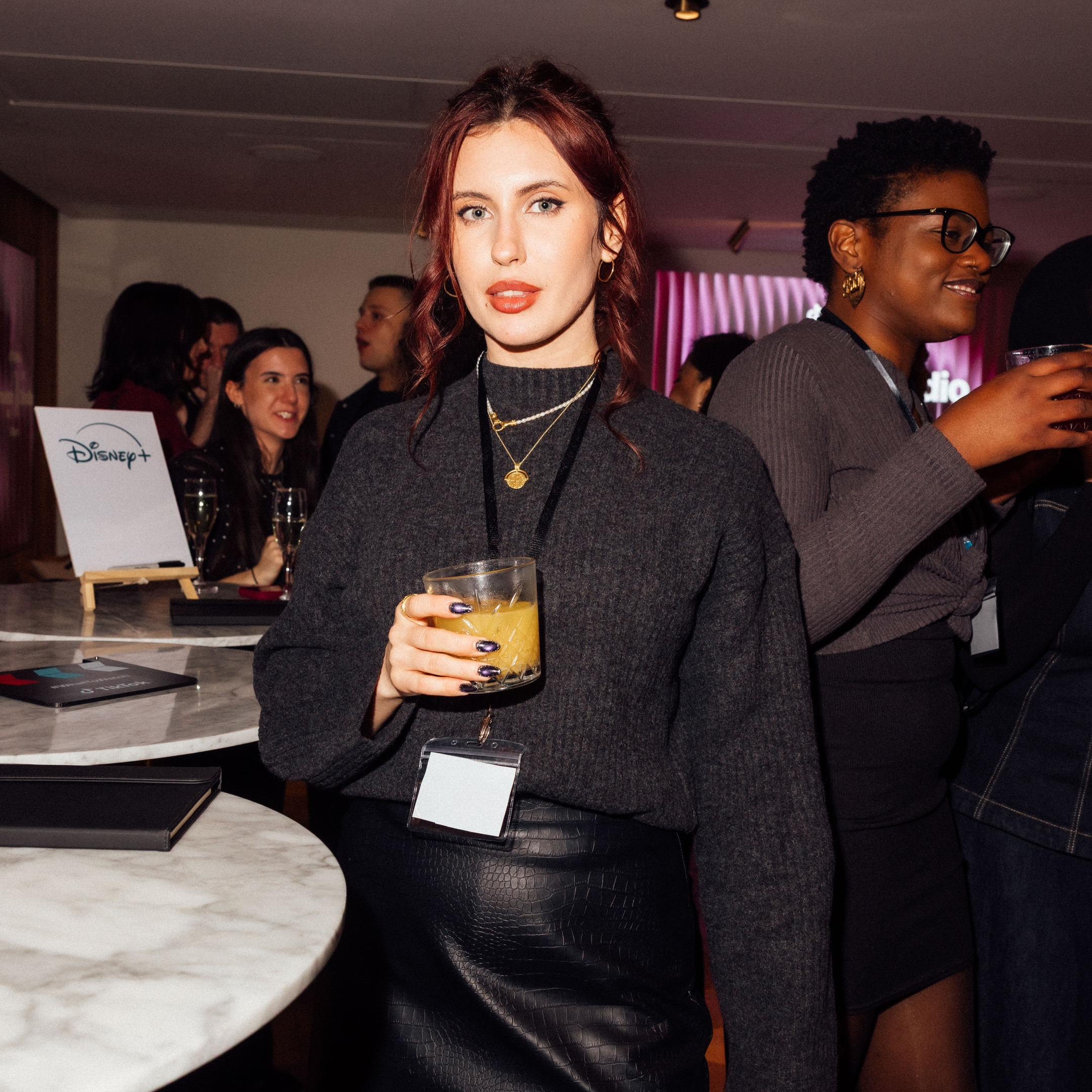 Woman with red hair holding a glass of yellow drink at a social event, with other people in the background and a Disney+ sign on the table.