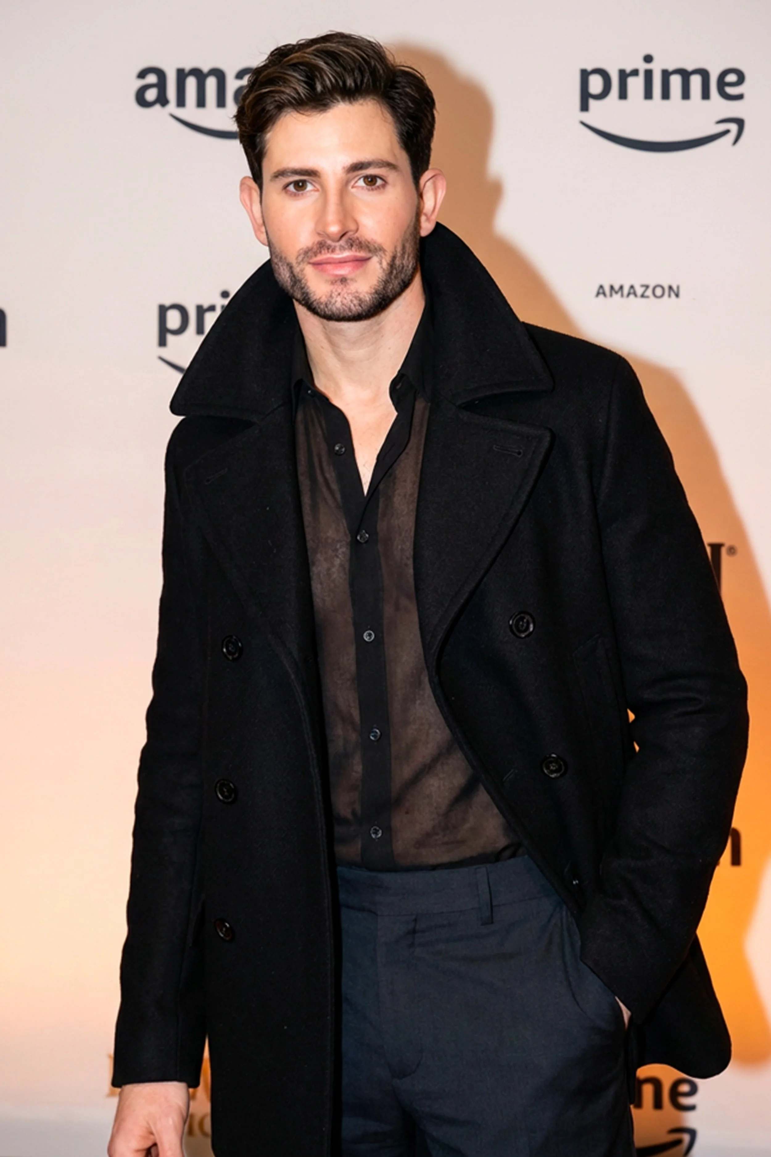 A man with dark hair and facial hair standing in front of a backdrop with Amazon and Prime logos, wearing a black coat over a dark shirt and dark pants.