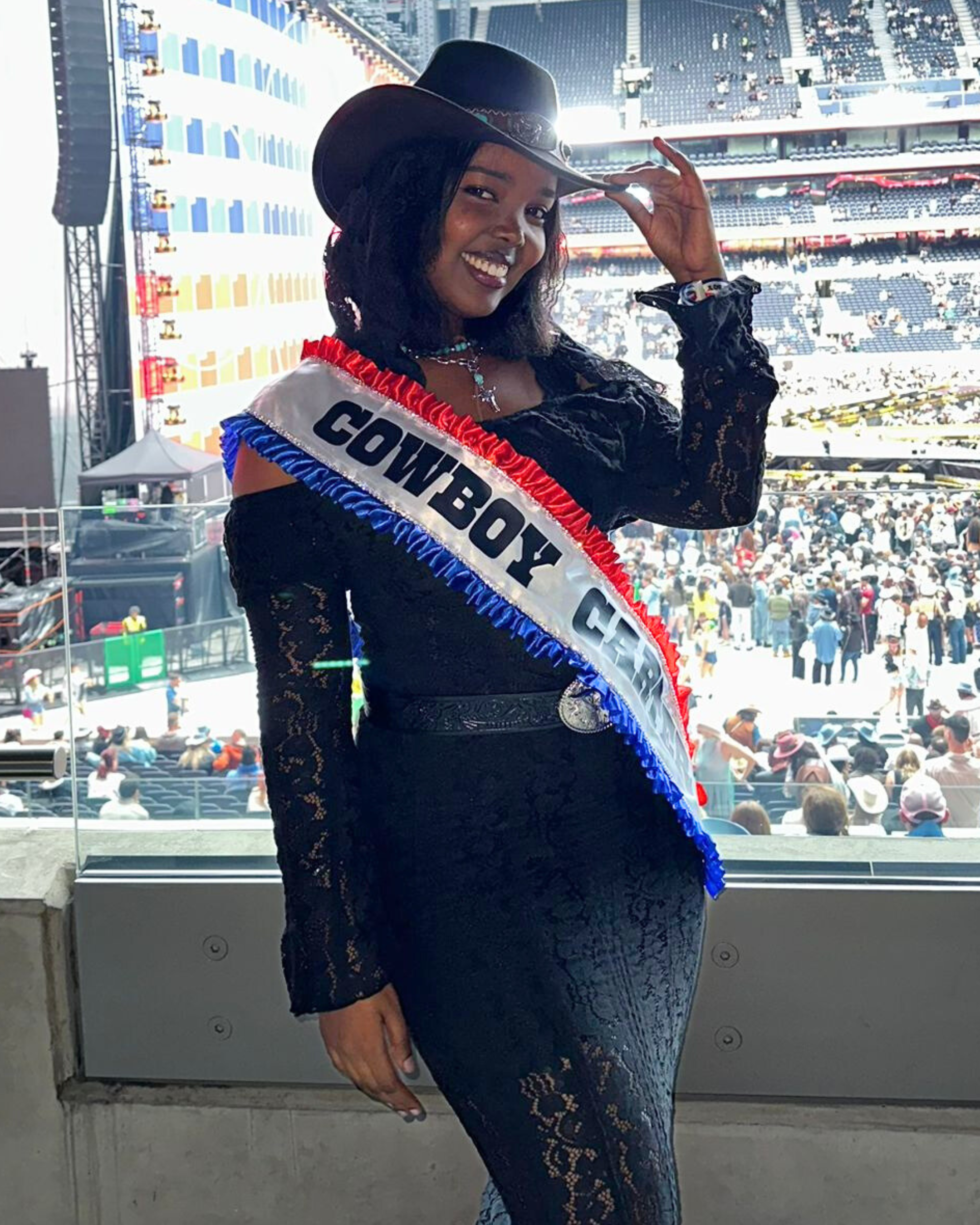 A woman in a black lace dress and a cowboy hat, wearing a sash that reads 'COWBOY GAL,' standing in a stadium with a crowd in the background.