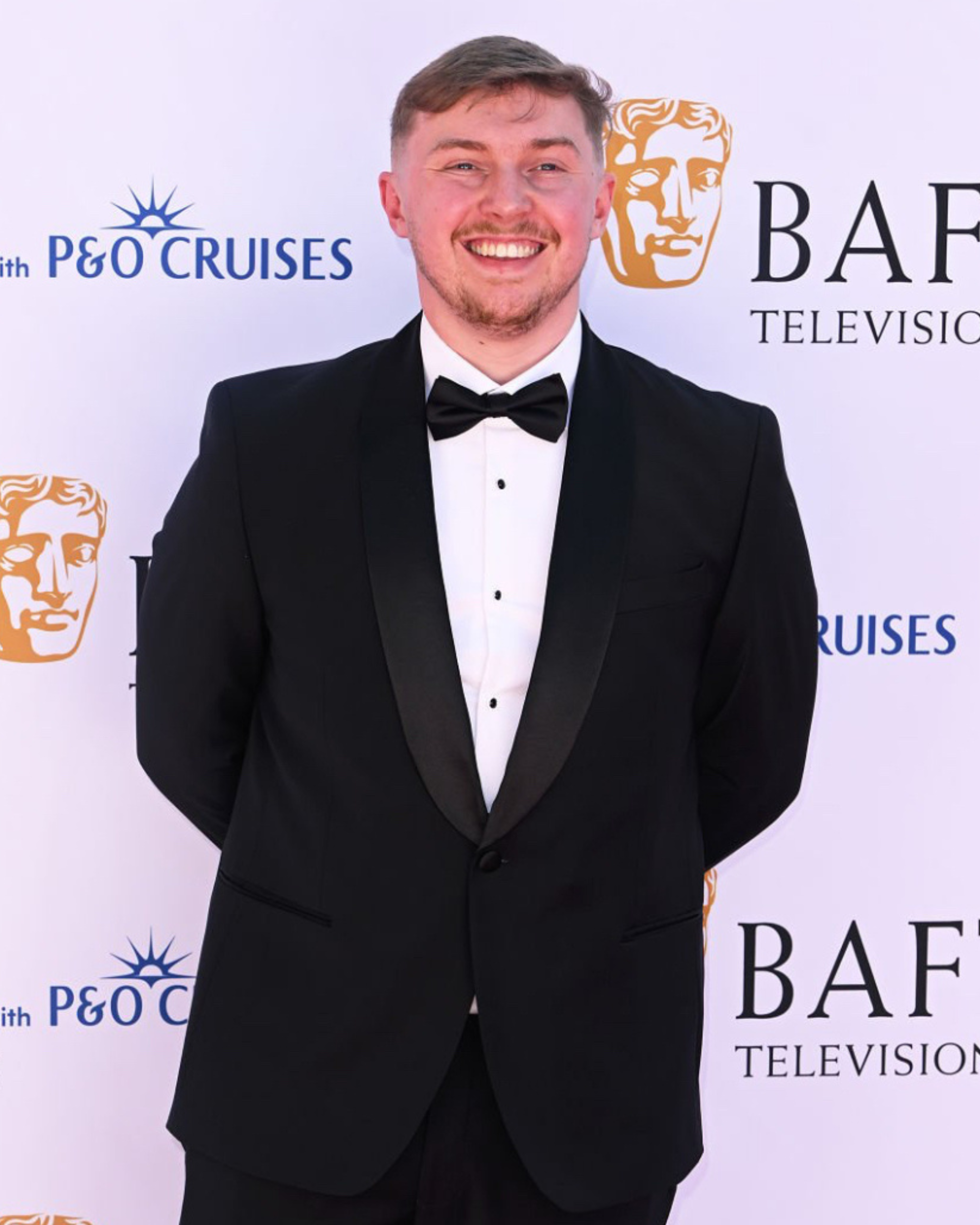 Man in a black tuxedo and bow tie smiling at a formal event, standing in front of a backdrop with logos and text.