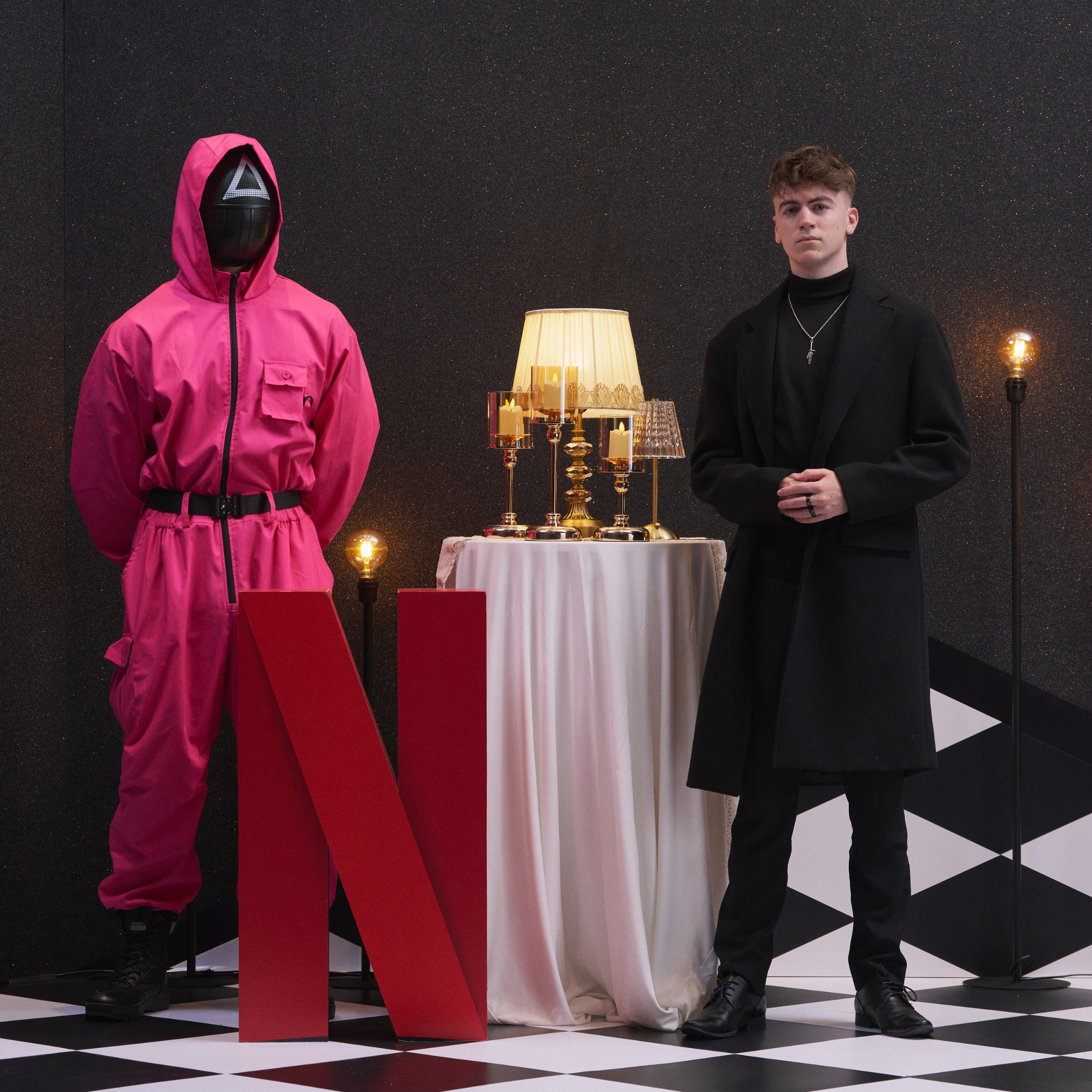 A person wearing a pink hooded jumpsuit with a mask standing next to a person dressed in black with a black long coat. They are in a room with a black glittery wall, checkered floor, and standing lamps. There is a white round table with gold and glass lamps on it, and a large red letter 'N' in front of the person in pink.