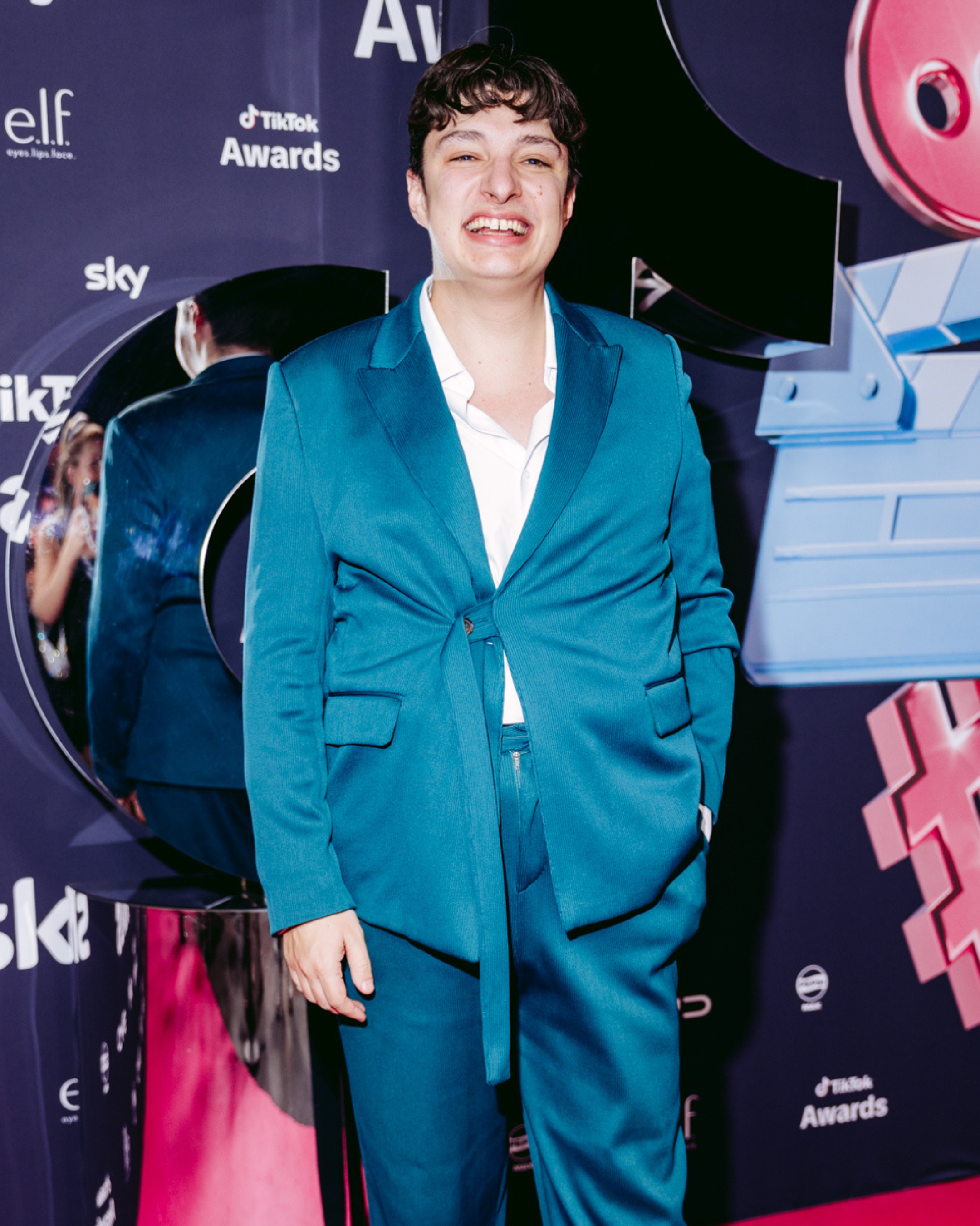 Young man in a blue suit standing in front of a backdrop with logos for TikTok, Sky, and Elf, smiling at an awards event.