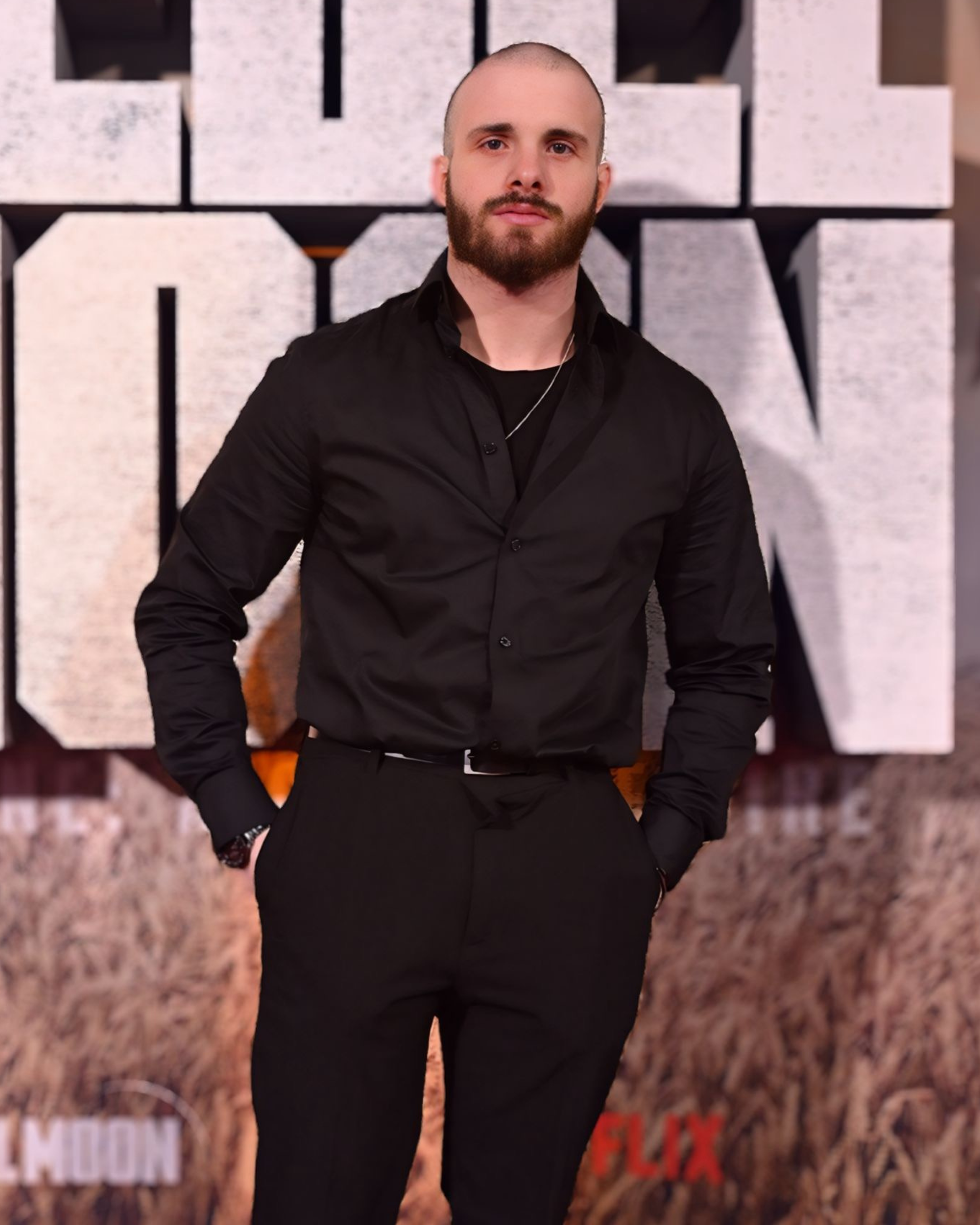 A man with a beard and shaved head, wearing a black shirt and black pants, standing with his hands in his pockets in front of a large textured sign with complex letterforms and the Netflix logo at the bottom.