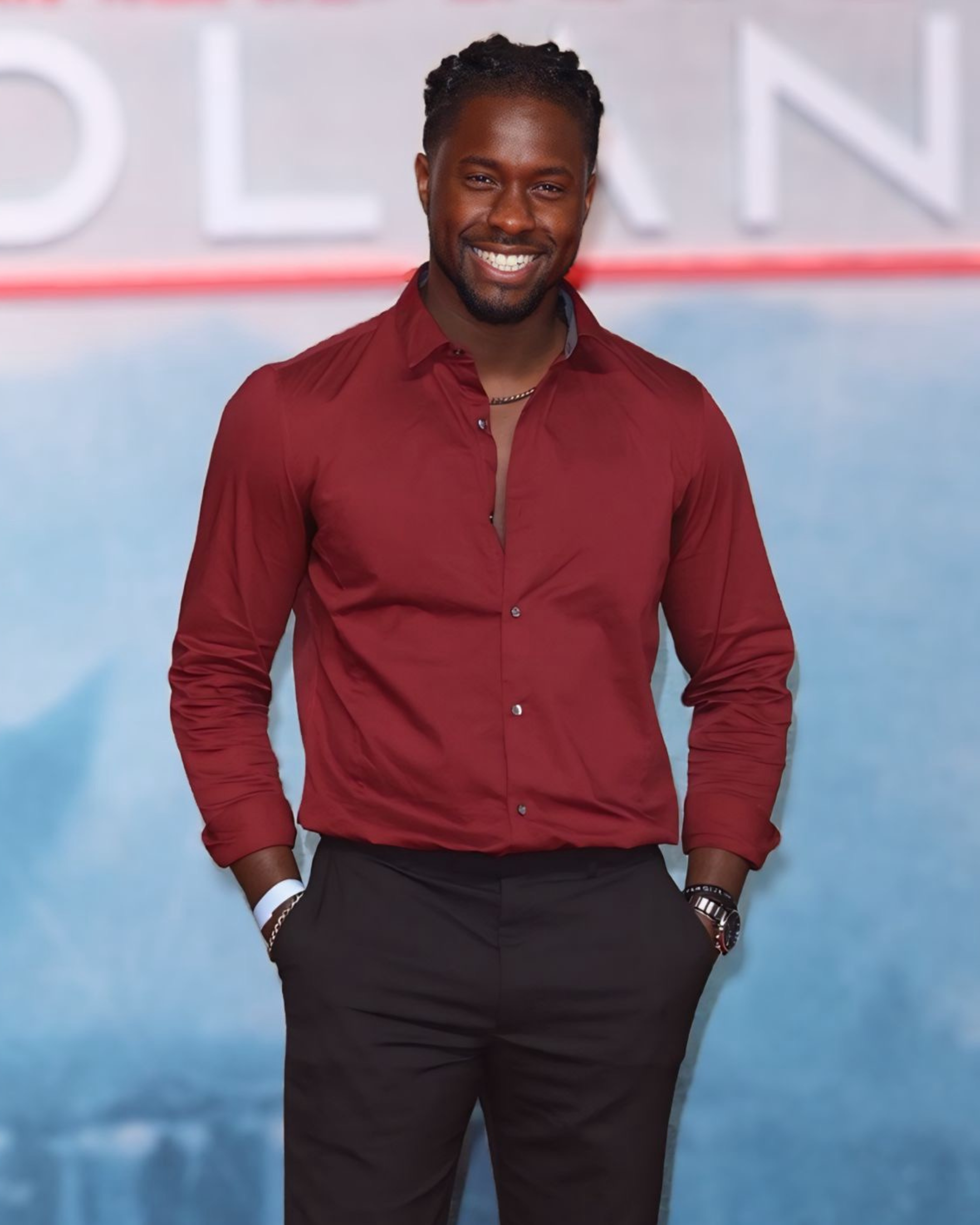 A smiling man with dark skin, wearing a red shirt and black pants, standing with his hands in his pockets.