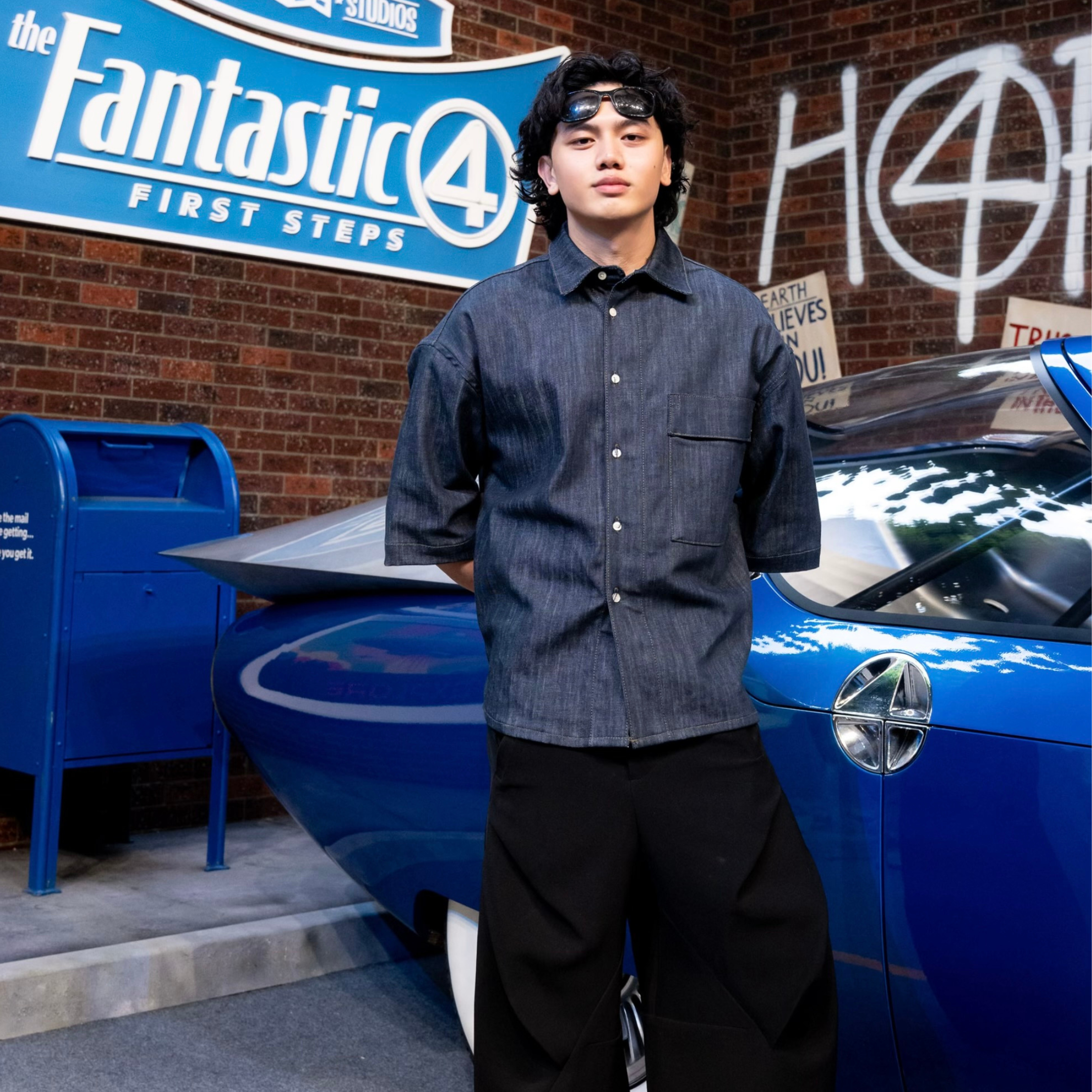 A young man with dark curly hair standing in front of a blue car and a blue mailbox, with a "Fantastic 4: First Steps" sign and graffiti on a brick wall behind him.
