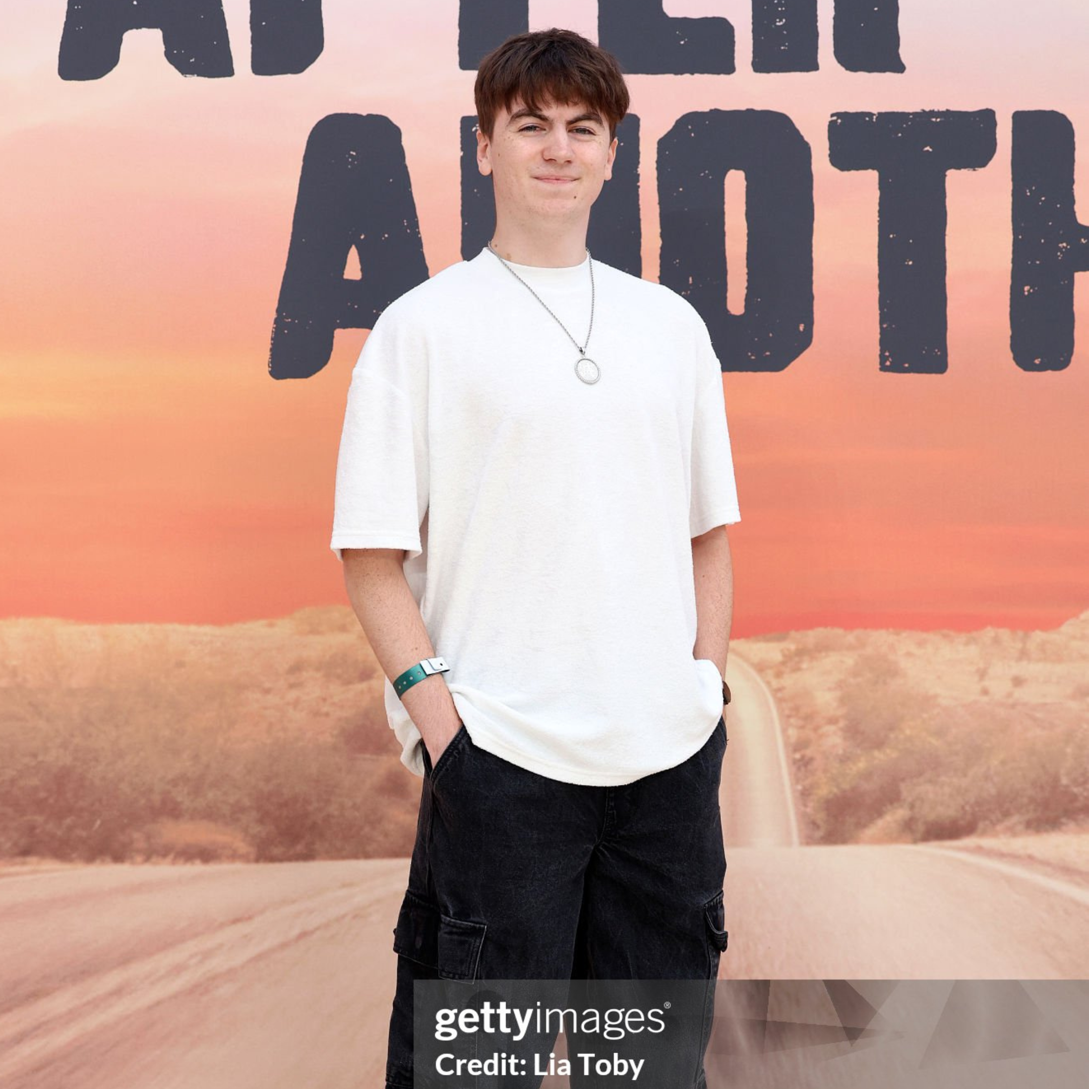 A young man standing outdoors at sunset with a not fully visible large sign behind him. He is wearing a white oversized t-shirt, black cargo shorts, a necklace, a watch, and a wristband, smiling slightly with hands in his pockets.