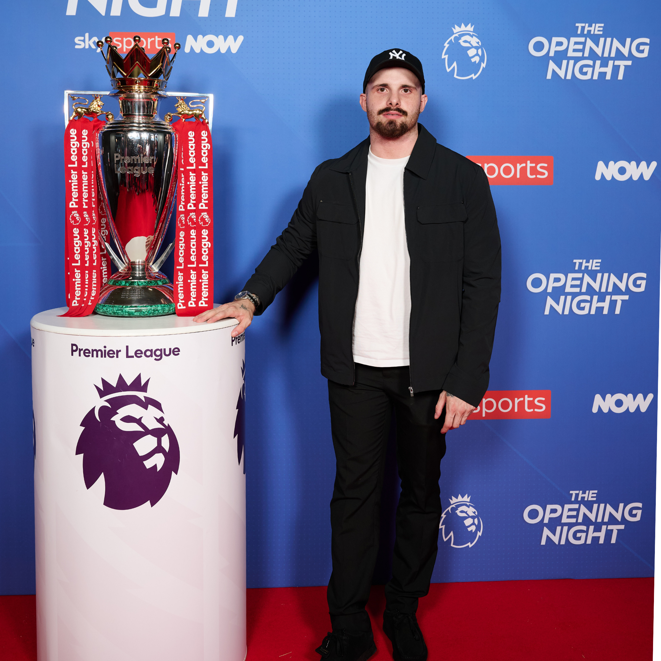 A man with a beard and mustache wearing a black jacket, white shirt, black pants, and a black New York Yankees cap standing next to the Premier League trophy on a white pedestal. The background features blue and red branding for the Premier League's opening night event with logos and text.