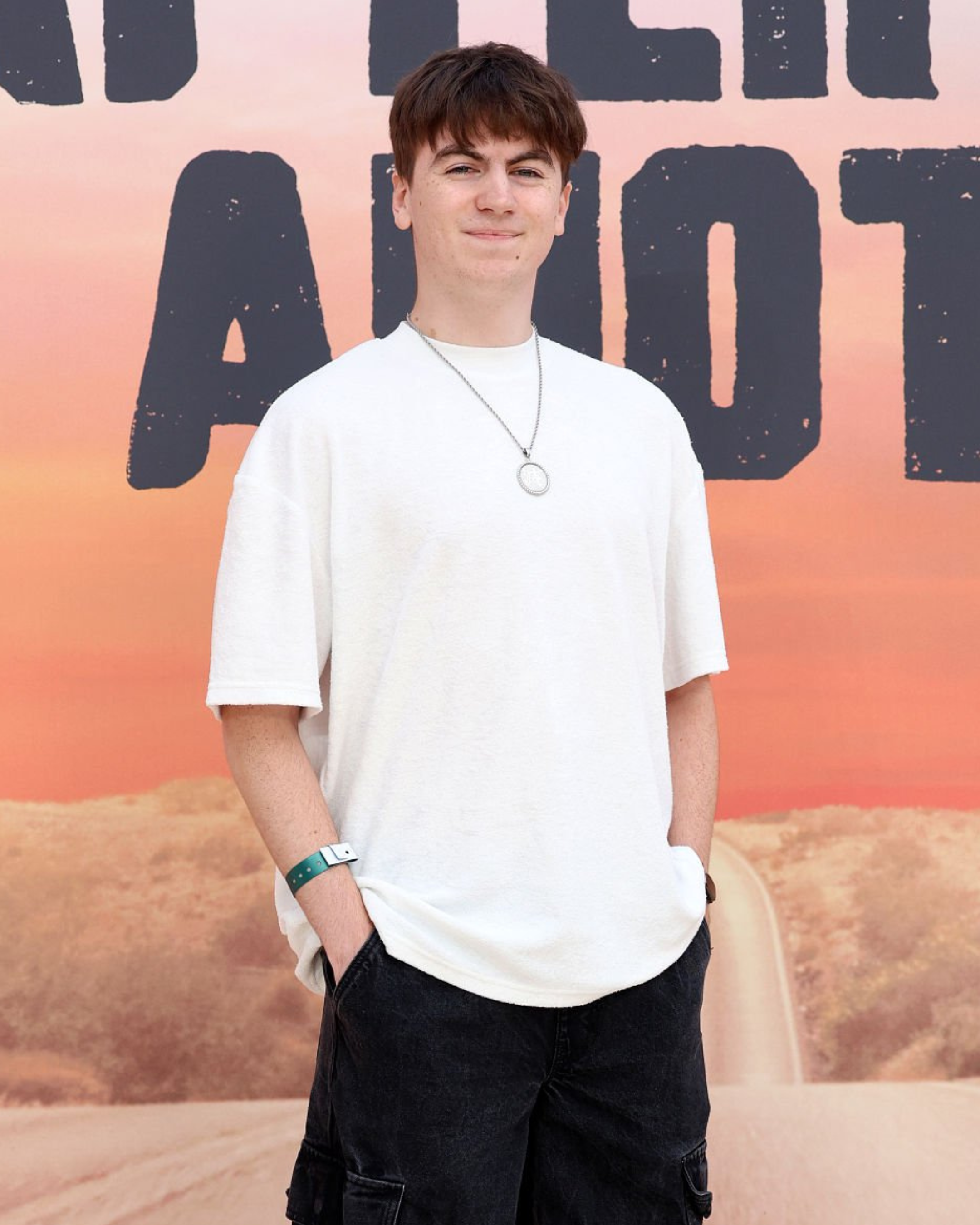 Teenage boy with brown hair and light skin standing in front of a sunset background with lettering. He wears a white oversized T-shirt, black shorts, a necklace, and a green wristband.