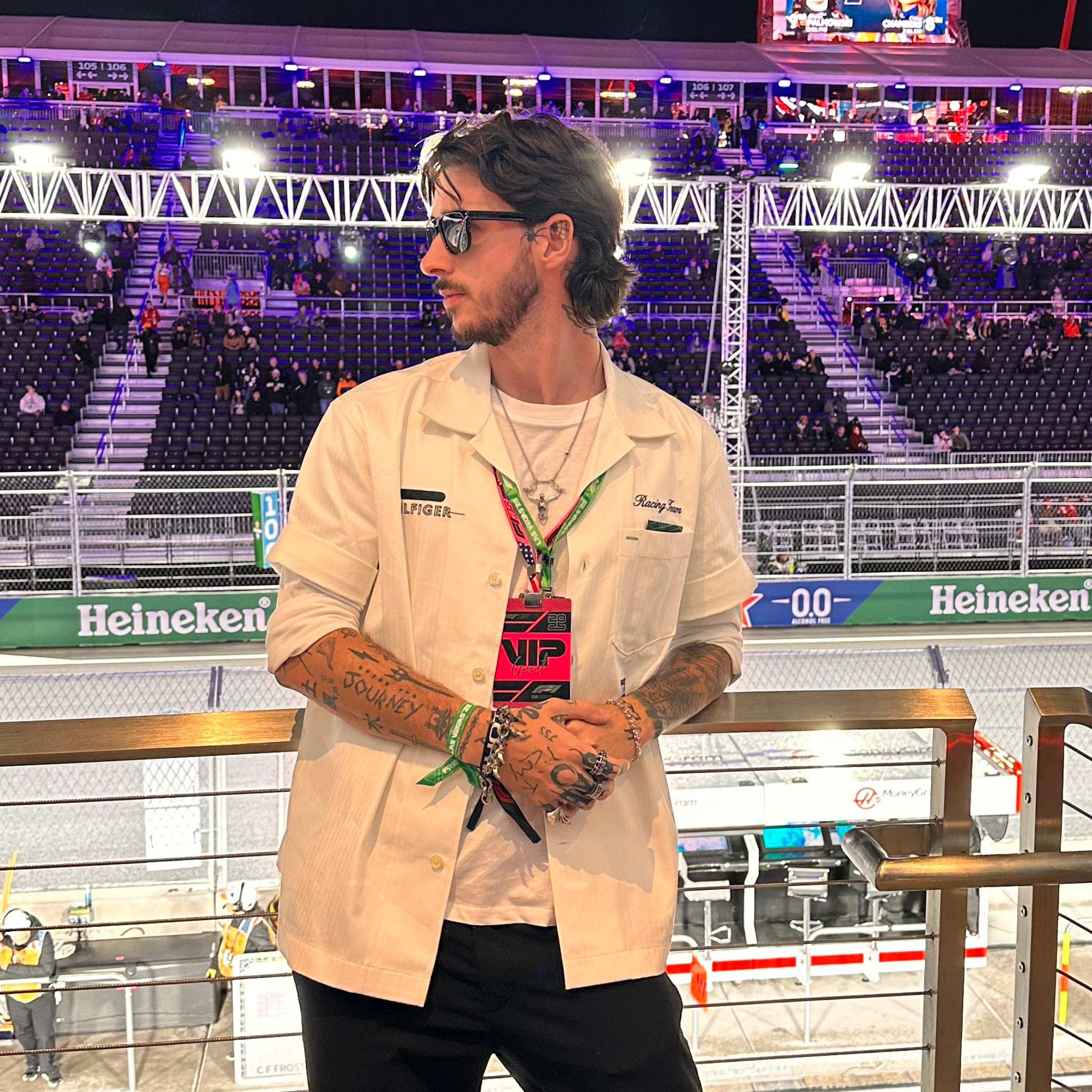 A young man with tattoos, dark hair, and sunglasses stands in front of a racing track at a nighttime event, wearing a beige racing shirt with patches, a white t-shirt underneath, multiple necklaces, and a pink VIP pass hanging around his neck.