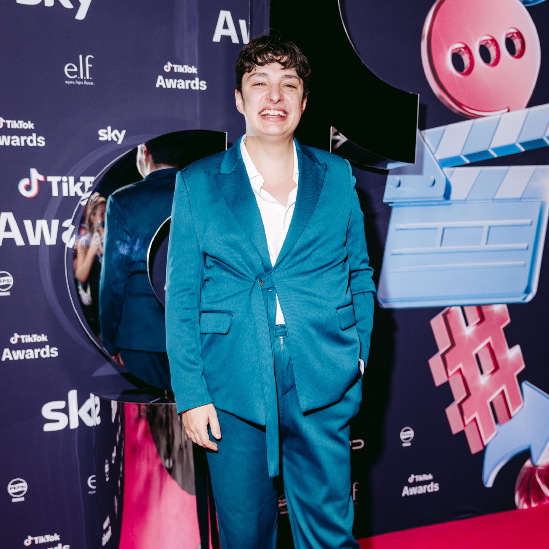 A person in a teal suit standing at the TikTok Awards event, smiling, with a backdrop featuring the TikTok logo, categories, and graphics.