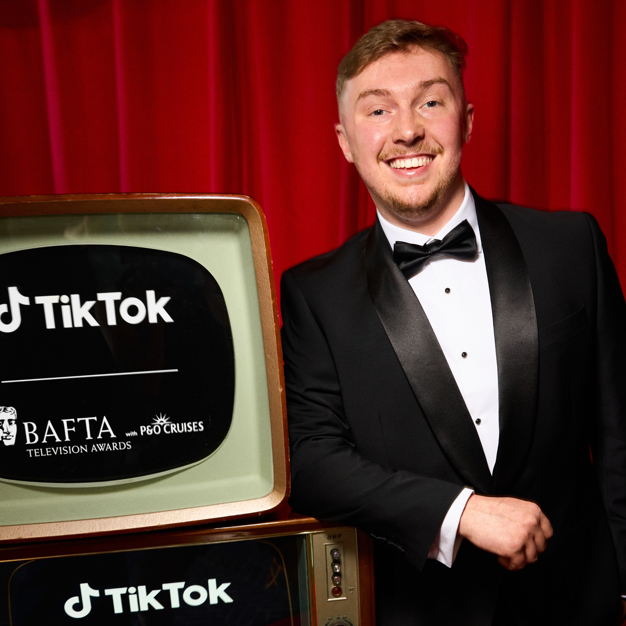 Man in black tuxedo smiling standing next to vintage television displaying TikTok and Bafta logos, red curtain background.