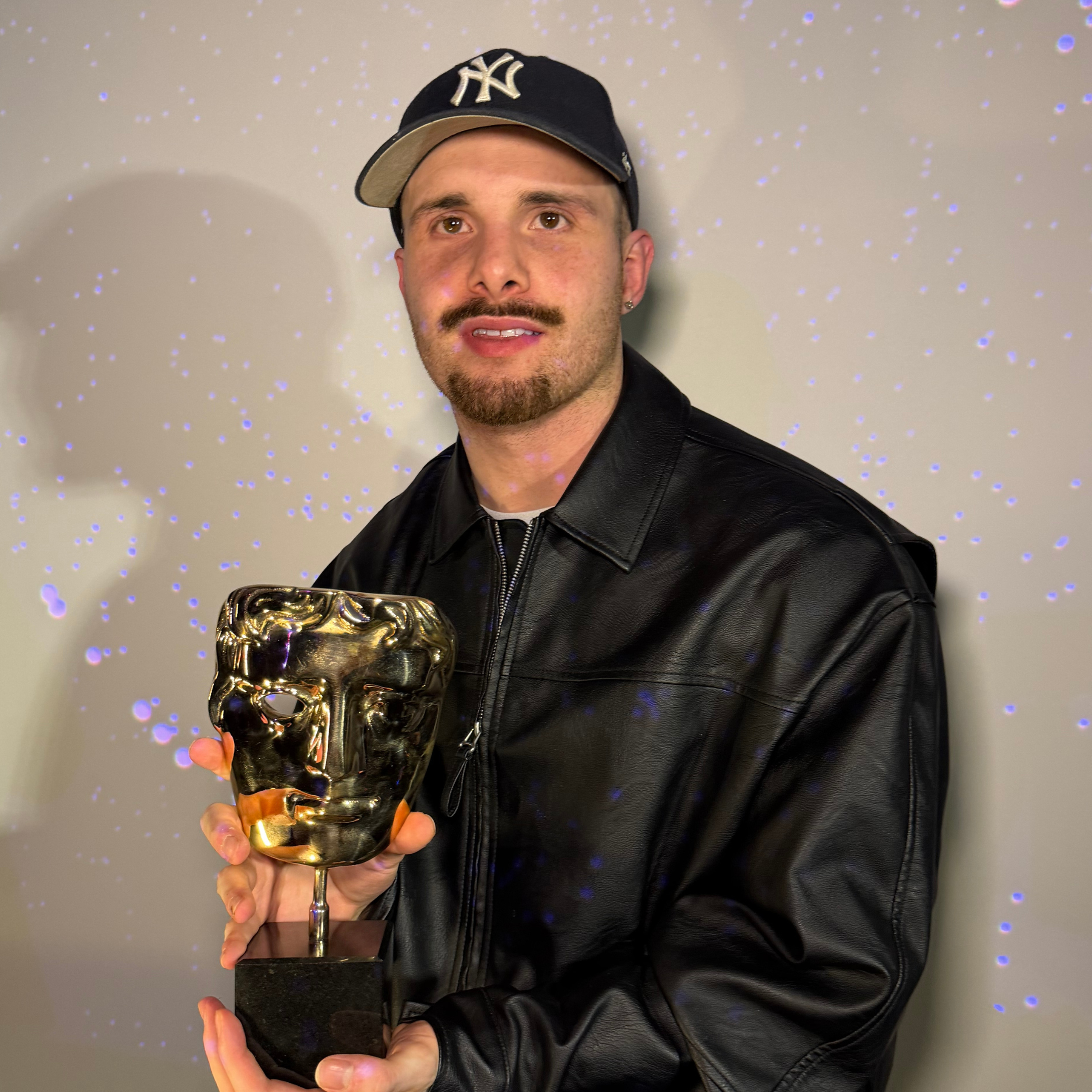 A man in a black leather jacket and a black New York Yankees cap holding a BAFTA award trophy, standing against a light-colored wall with purple and blue light spots.