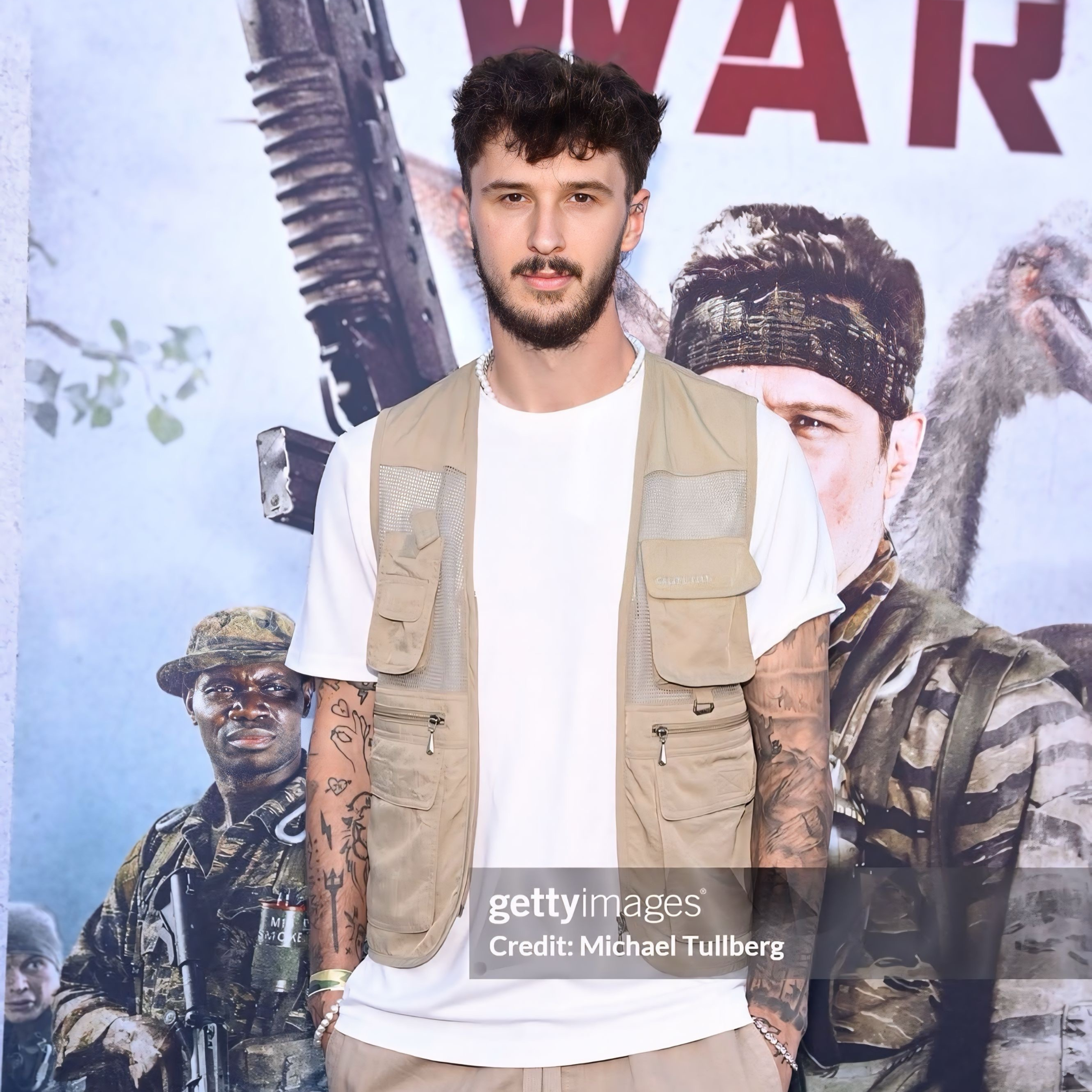 A young man with curly brown hair and a beard, wearing a beige tactical vest over a white t-shirt, standing in front of a promotional poster for the movie 'War' featuring soldiers in military gear and weapons.