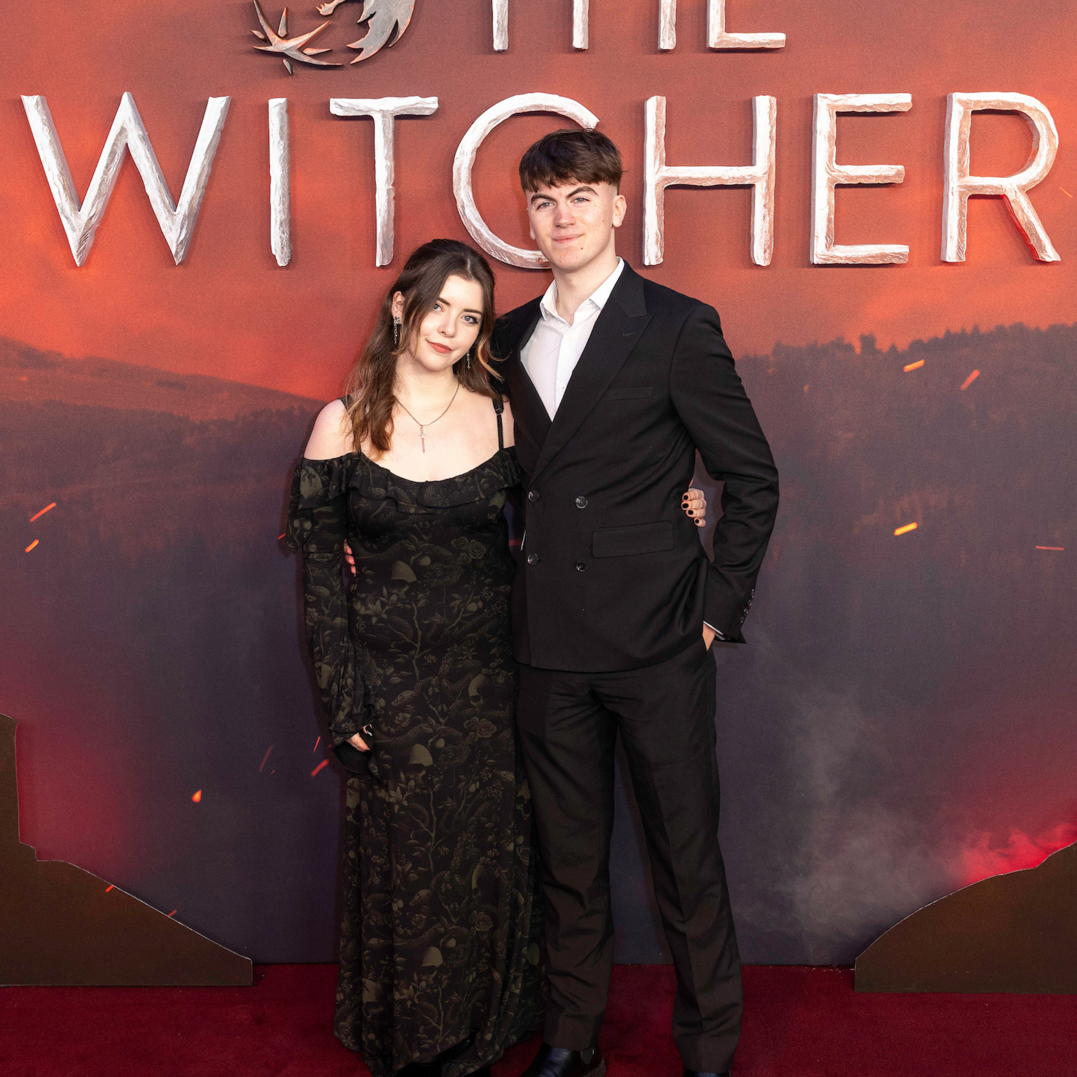 A young woman and a young man dressed in formal attire standing together at an event with a backdrop labeled 'The Witcher'. The woman is wearing a black, patterned dress with off-the-shoulder sleeves, and the man is in a black suit with a white shirt. They are smiling at the camera.