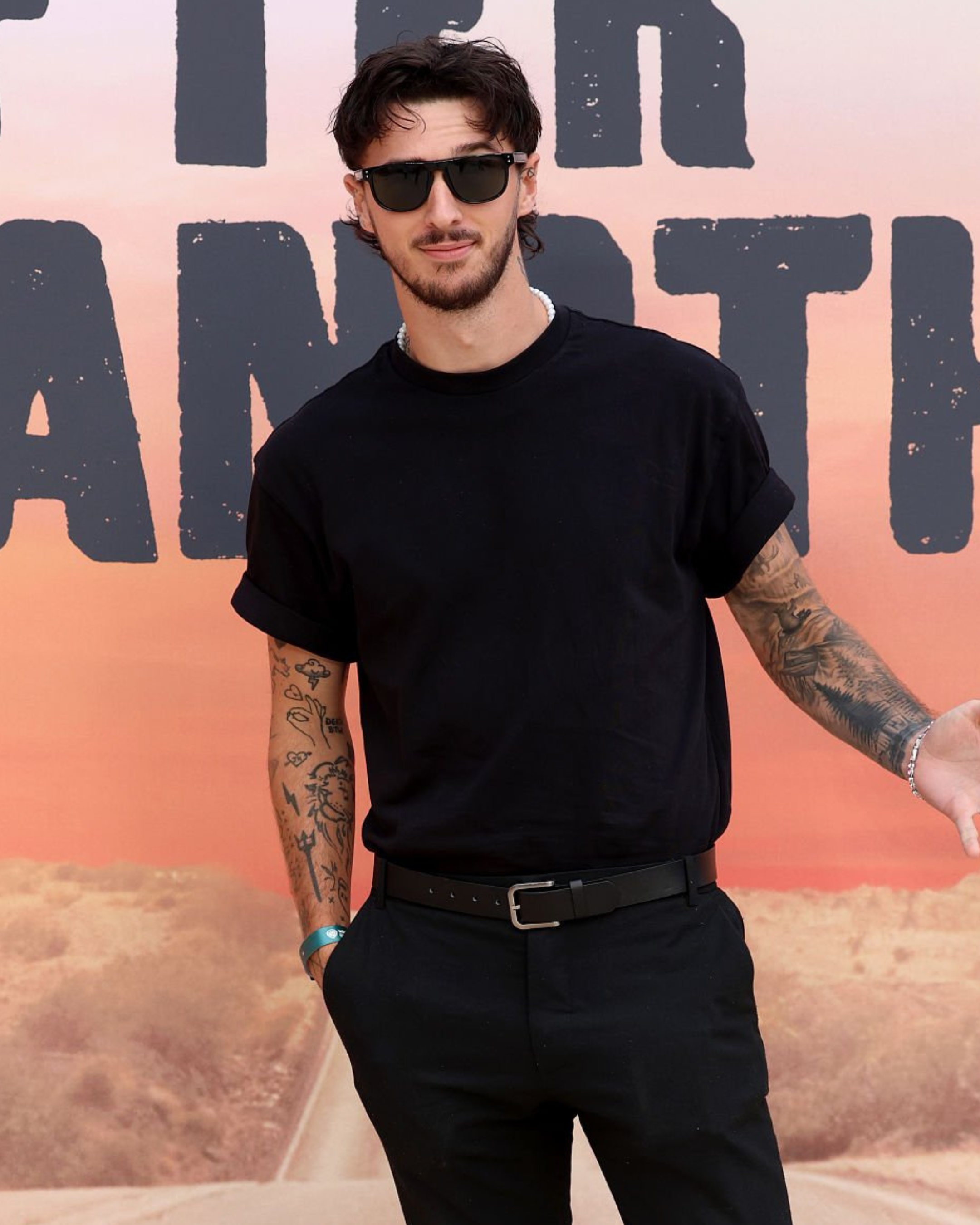 A man with dark hair, beard, and sunglasses wearing a black t-shirt with rolled sleeves, black pants, and a black belt, standing in front of a desert background with large text.