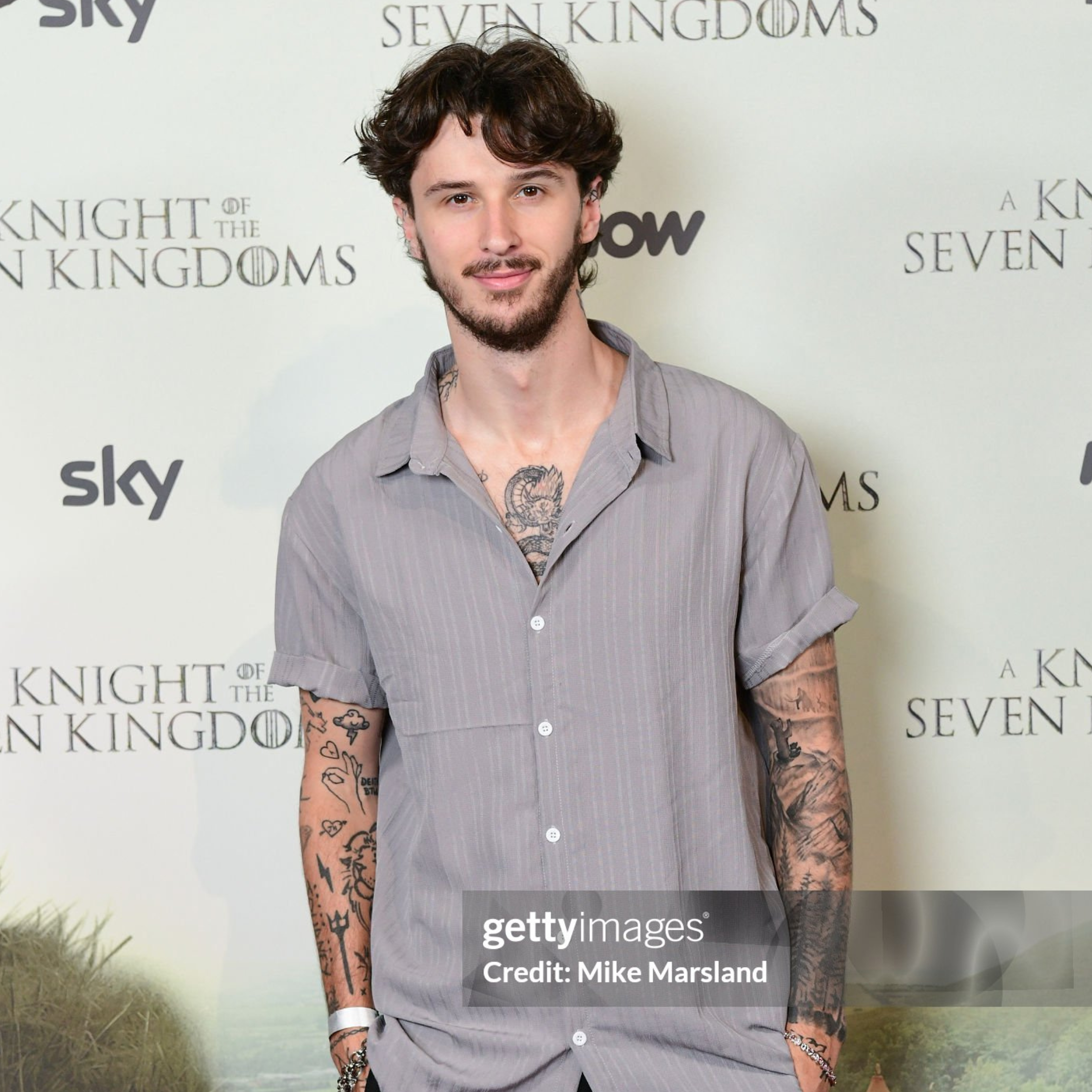 A young man with dark brown curly hair, beard, and tattoos on his arms and chest, standing in front of a backdrop with text for television shows and sky. He is wearing a light gray, short-sleeved, collared shirt and smiling slightly.