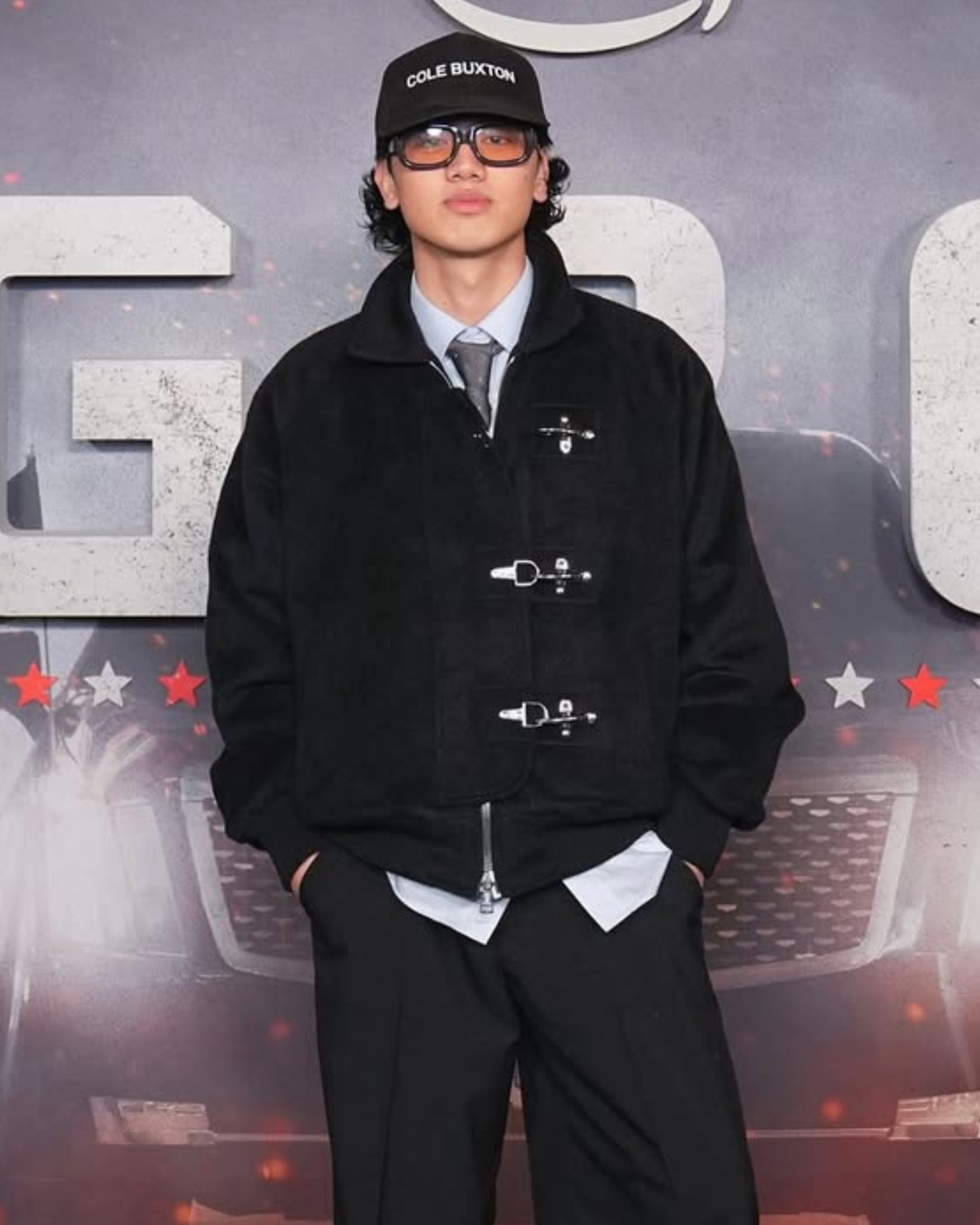 Young man wearing a black cap with white text, sunglasses, a black jacket with toggle closures, a gray shirt, and black pants standing in front of a backdrop with partial cars and stars.